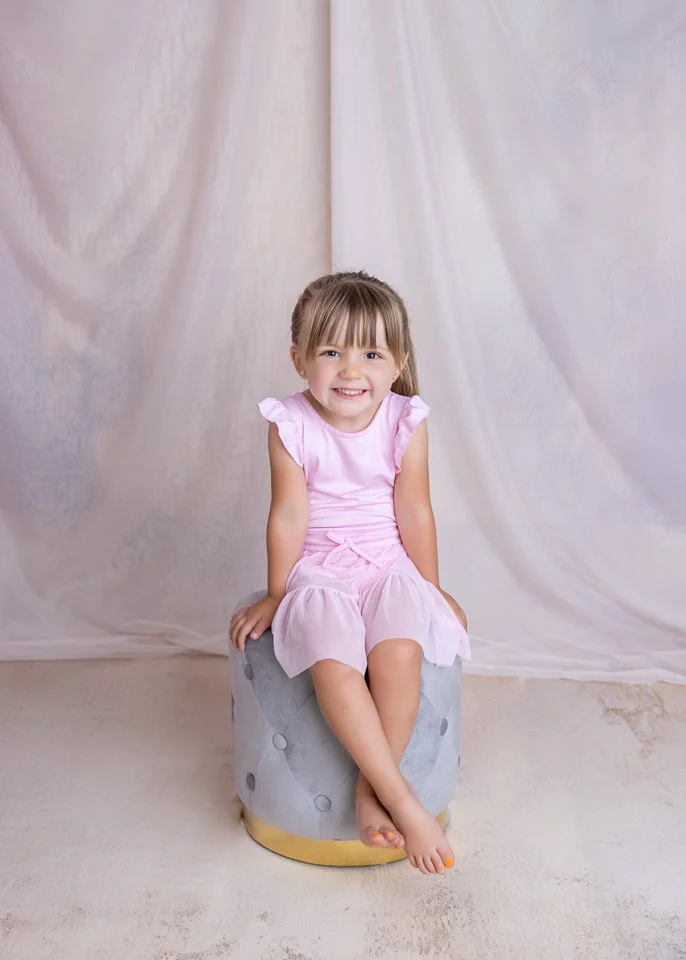 A young girl in a pink dress sitting on a gray, tufted ottoman, smiling at the camera, with a light-colored backdrop.
