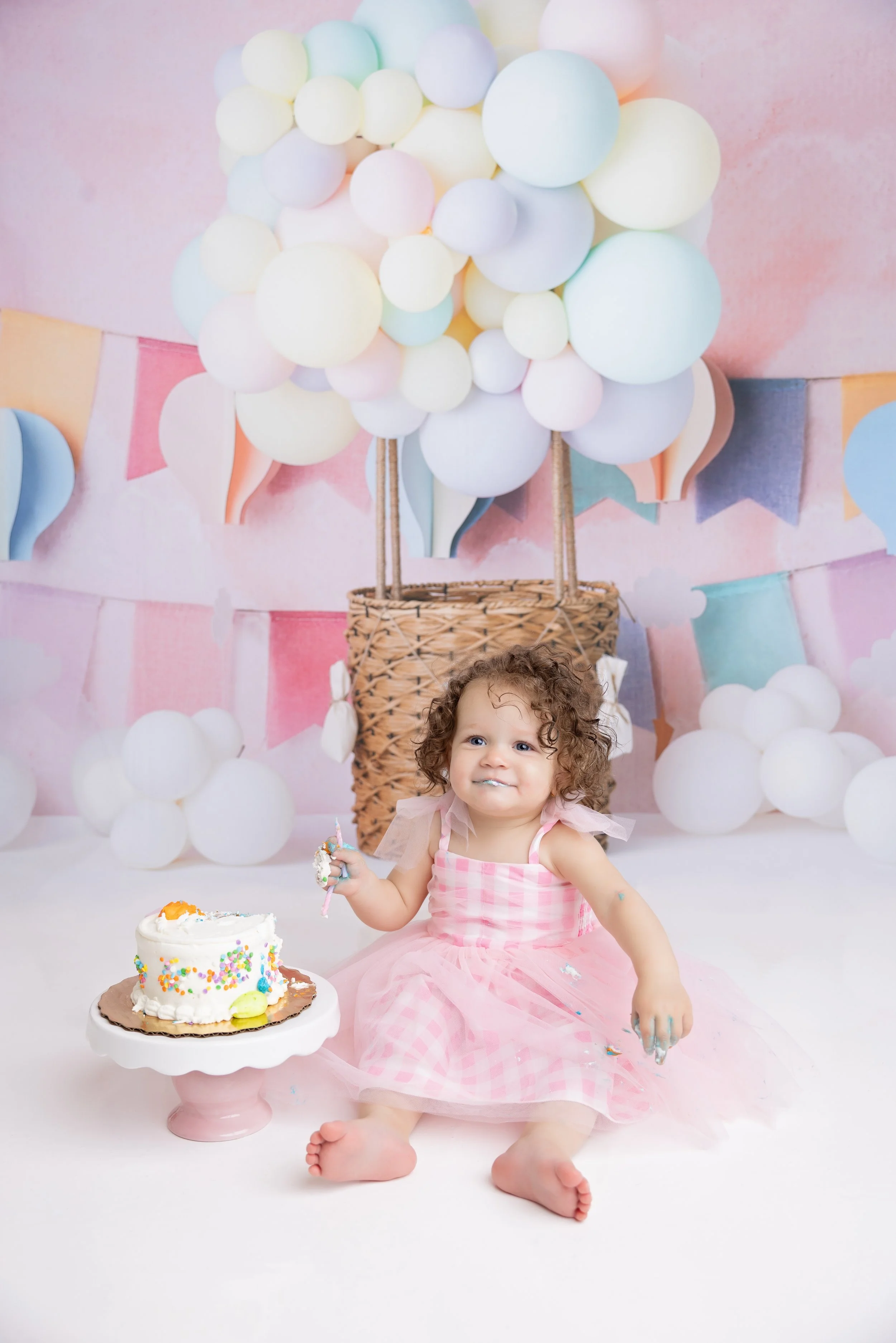 A young girl in a pink dress is sitting on the floor with a partially eaten birthday cake in front of her, set against a pastel-themed backdrop with balloons and bunting.