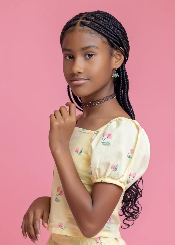 A young girl with braided hair wearing a yellow floral dress, a choker necklace, and earrings, posing against a pink background.