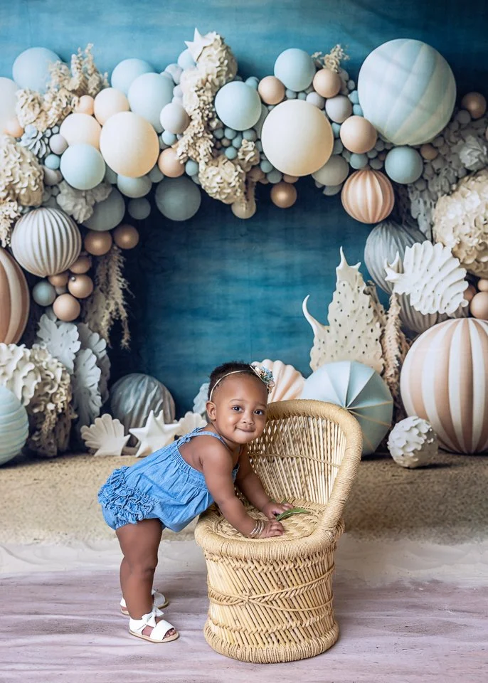 A young girl in a blue dress and white sandals leaning on a wicker chair, in front of a pastel-colored balloon and seashell-themed backdrop.
