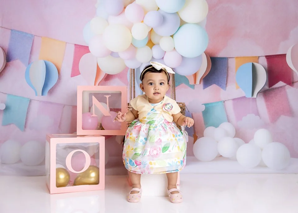 A baby girl sitting in front of a pastel balloon and banner backdrop for a celebration, surrounded by pastel and gold decorative boxes with balloons inside.