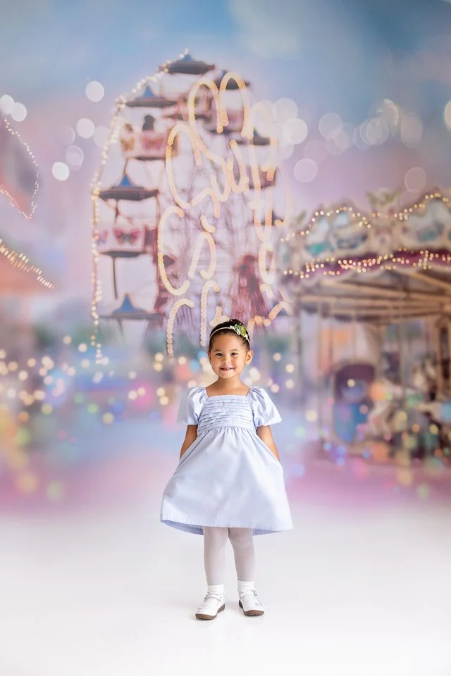 Young girl in white dress standing in front of colorful blurry carnival background with Ferris wheel and bright lights.