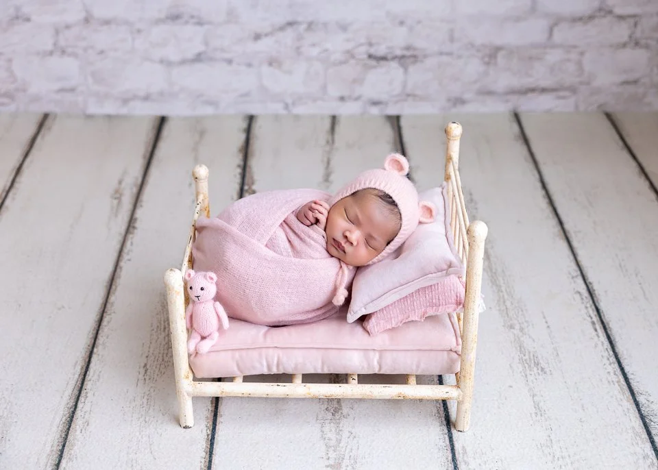 Newborn baby sleeping on a small white bed, wrapped in pink cloth, wearing a pink hat with bear ears, with a matching teddy bear and pillow, on a light wooden floorboard with a white brick wall background.