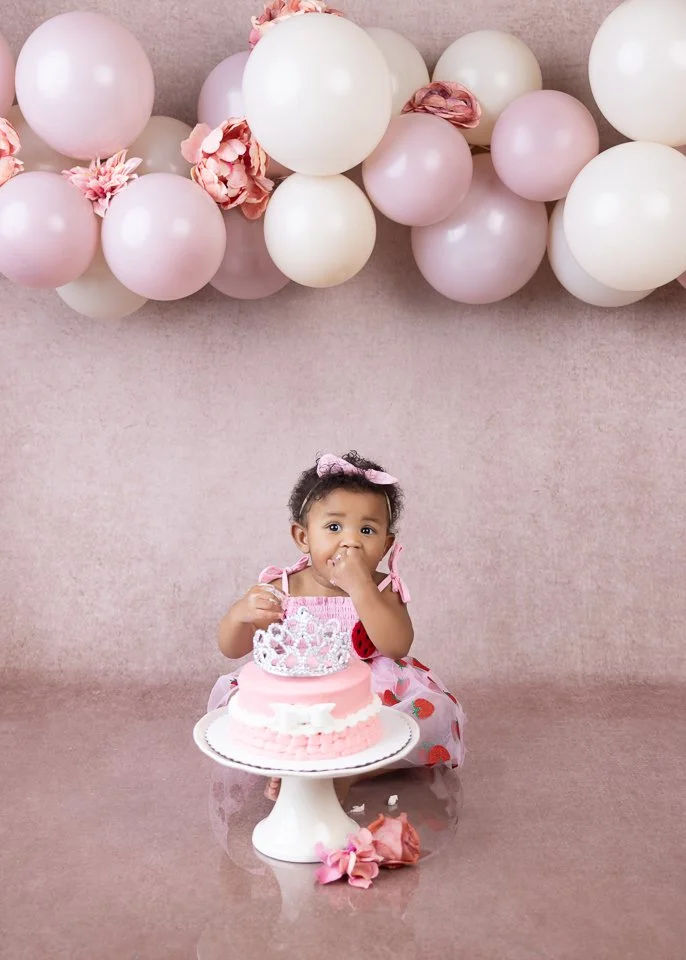 A young girl in a pink dress with a bow on her head sitting behind a pink birthday cake with white and pink decorations, in front of a backdrop decorated with pink and white balloons and flowers.
