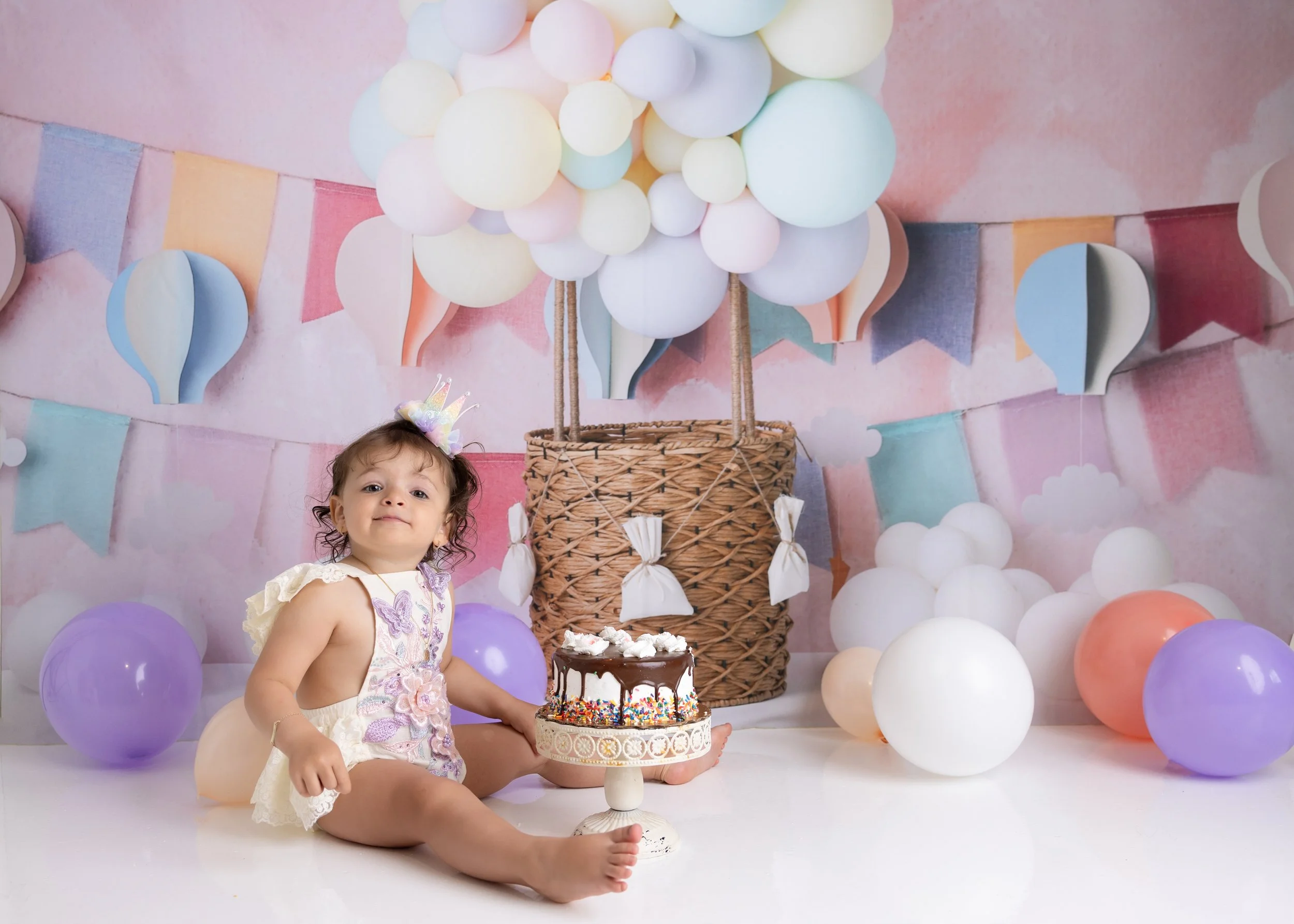 A young girl celebrating her birthday with a pastel-colored balloon backdrop, wearing a party hat, sitting on the floor, with a decorated cake in front of her.
