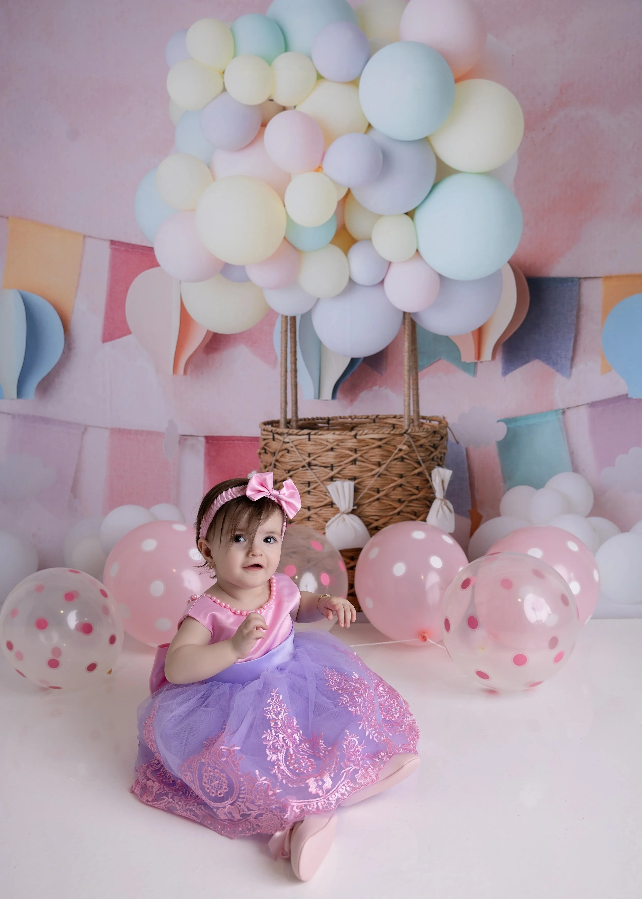 A young girl sitting on the floor at a birthday party, wearing a pink and purple dress with a pink bow headband, surrounded by pink branded balloons and a backdrop with pastel-colored balloons and bunting.