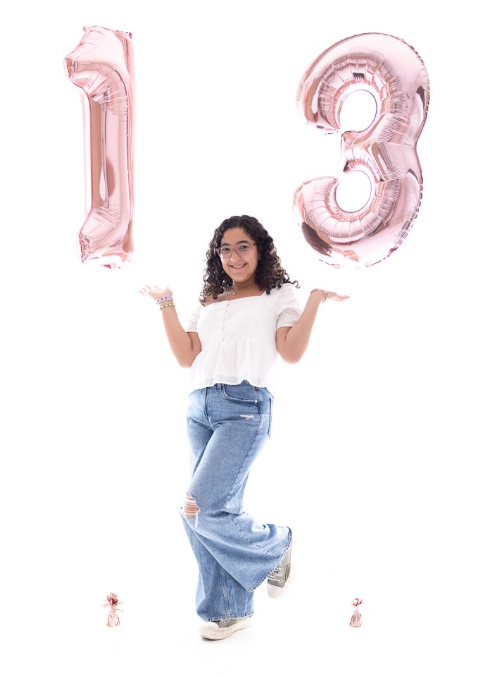 Young woman with curly hair and glasses holding pink number balloons '1' and '3' celebrating a 13th birthday, standing against a white background.
