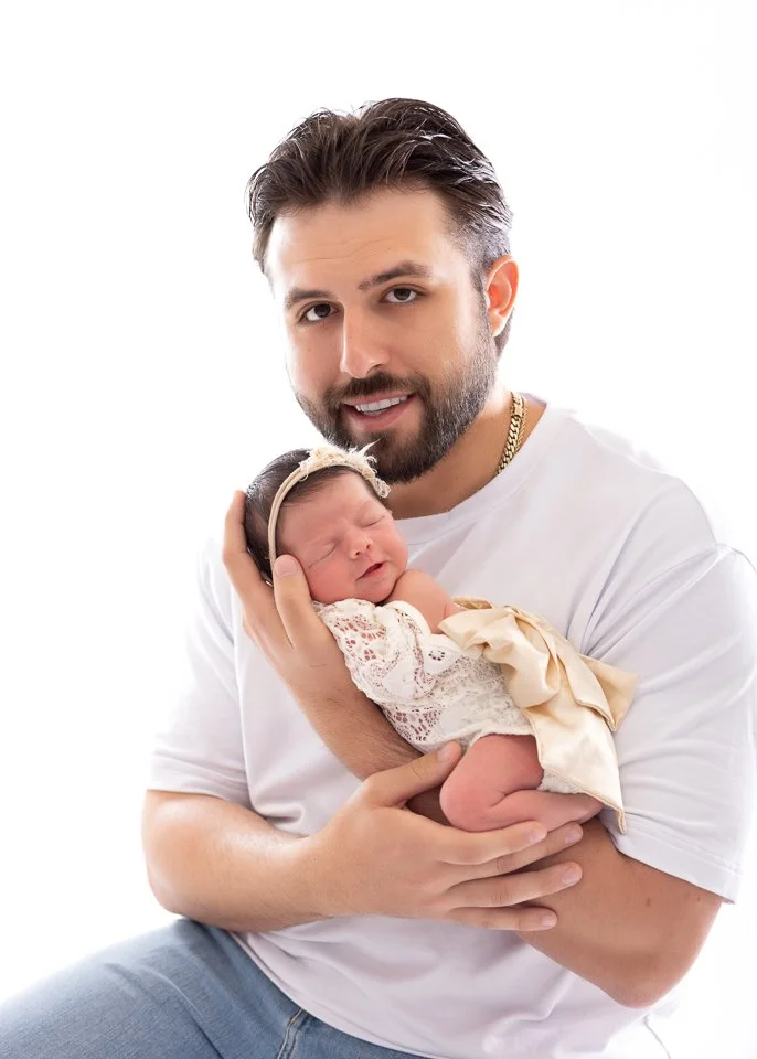 A man holding a newborn baby girl dressed in cream-colored lace and satin clothing with a bow, against a plain white background.