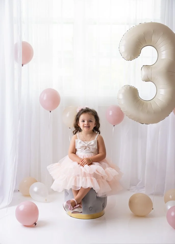 A young girl in a pink tutu dress sitting on a small styled stool surrounded by pinks and beige balloons, with a large silver number three balloon indicating her third birthday celebration.