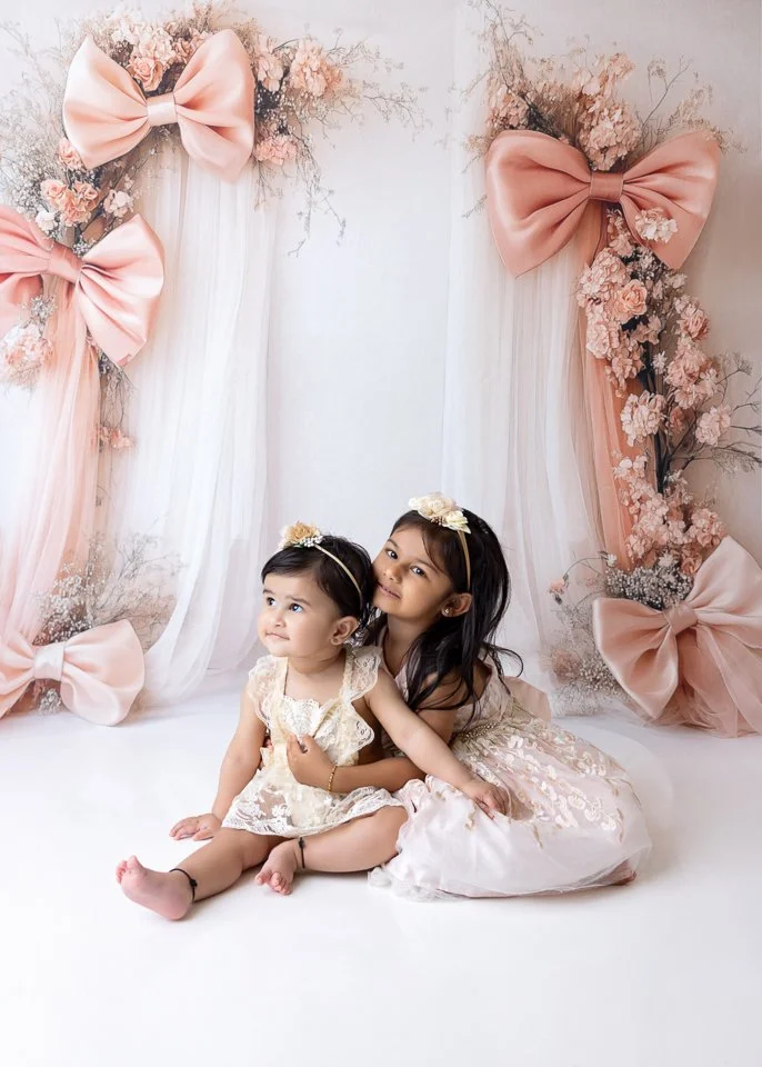 Two young girls sitting against a floral backdrop with pink bows and draped fabric, dressed in elegant dresses and floral headbands.