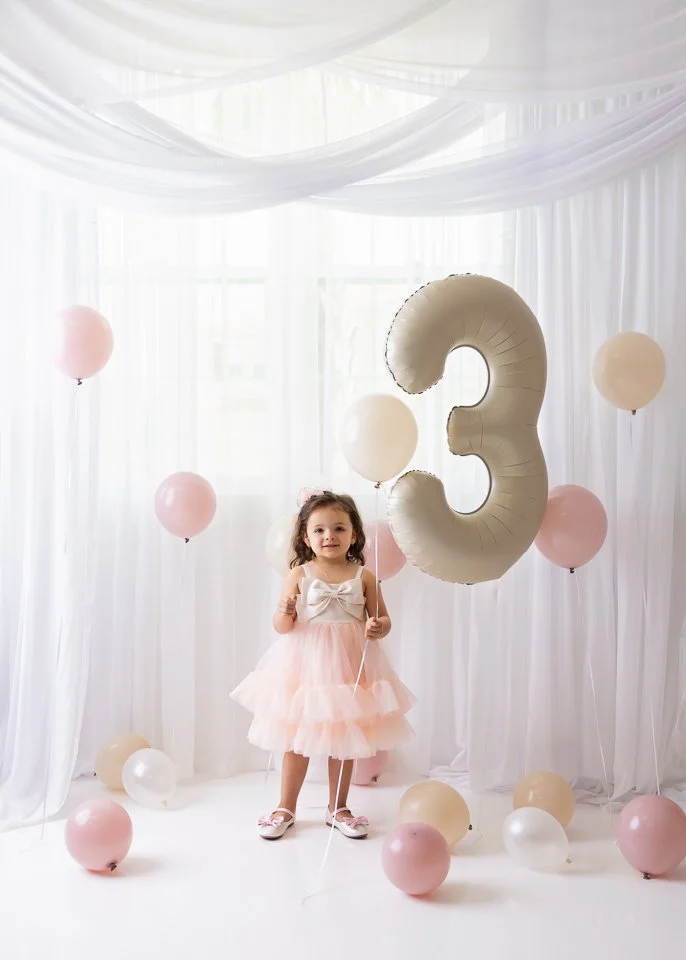 A young girl in a light pink dress holding balloons, standing in a decorated room with white drapes and a large balloon shaped as the number 3, celebrating her third birthday.