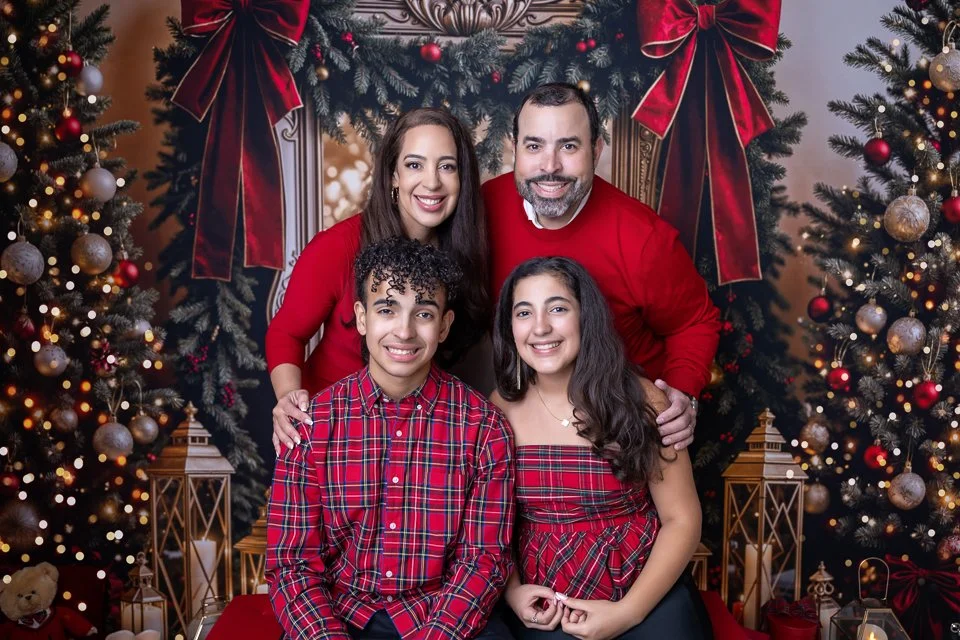 Family of five in Christmas sweaters sitting and standing in front of decorated Christmas trees with ornaments and lights.
