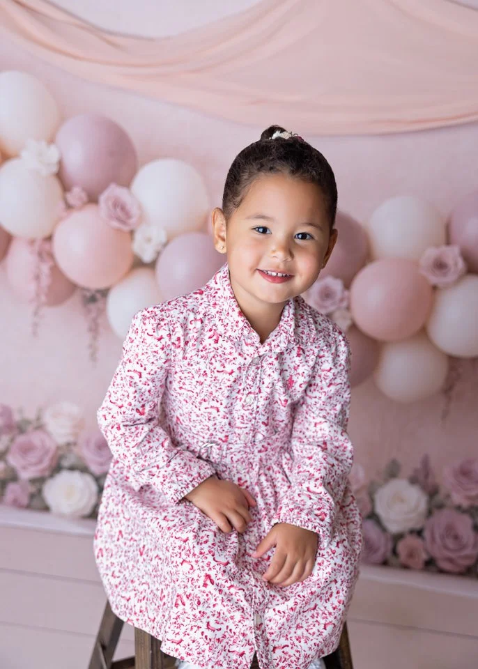 A young girl with a high ponytail, wearing a floral dress, sitting on a stool, smiling at the camera, with pink and white balloons and flowers in the background.
