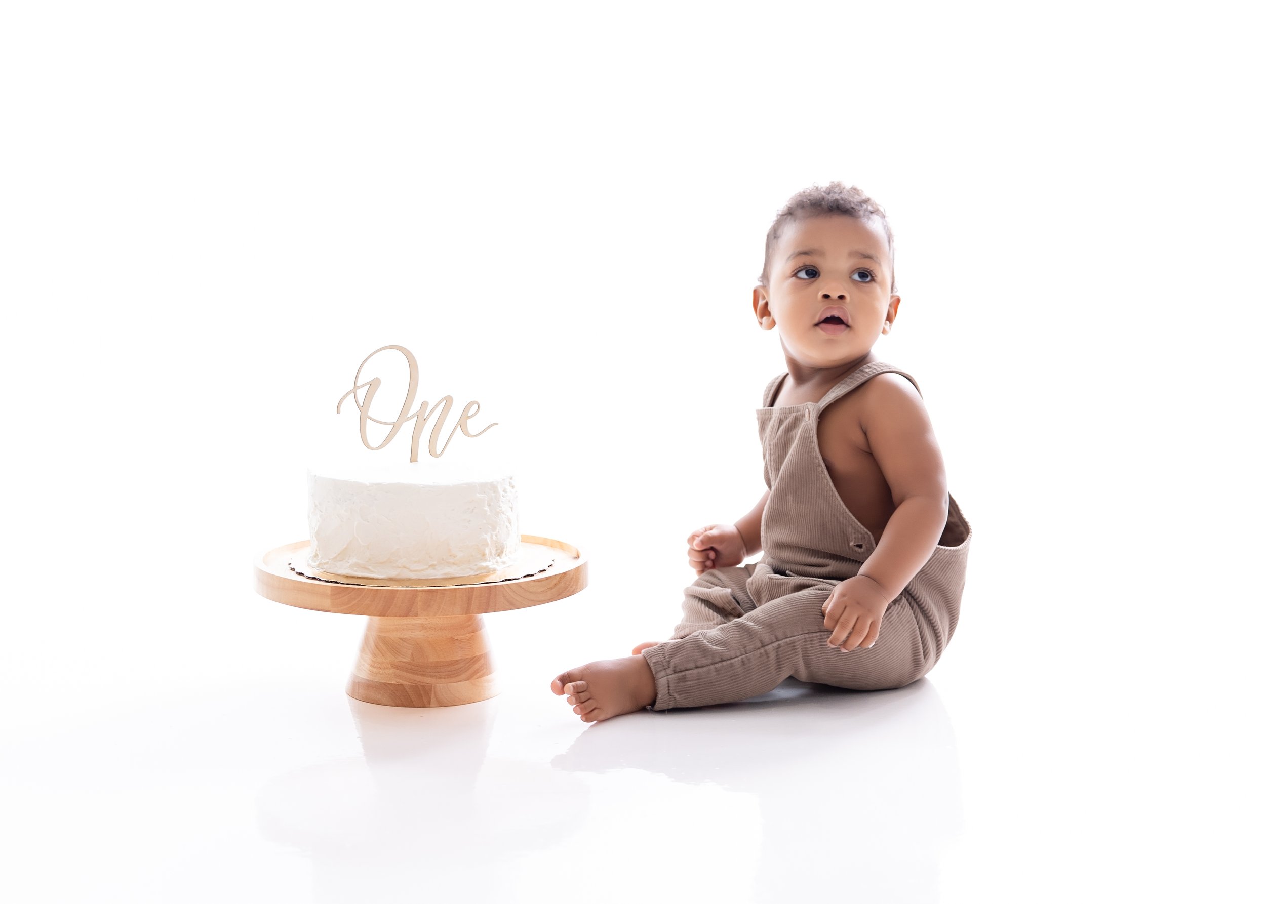 Cute toddler sitting on the floor next to a white birthday cake with a gold 'One' topper, on a white background.