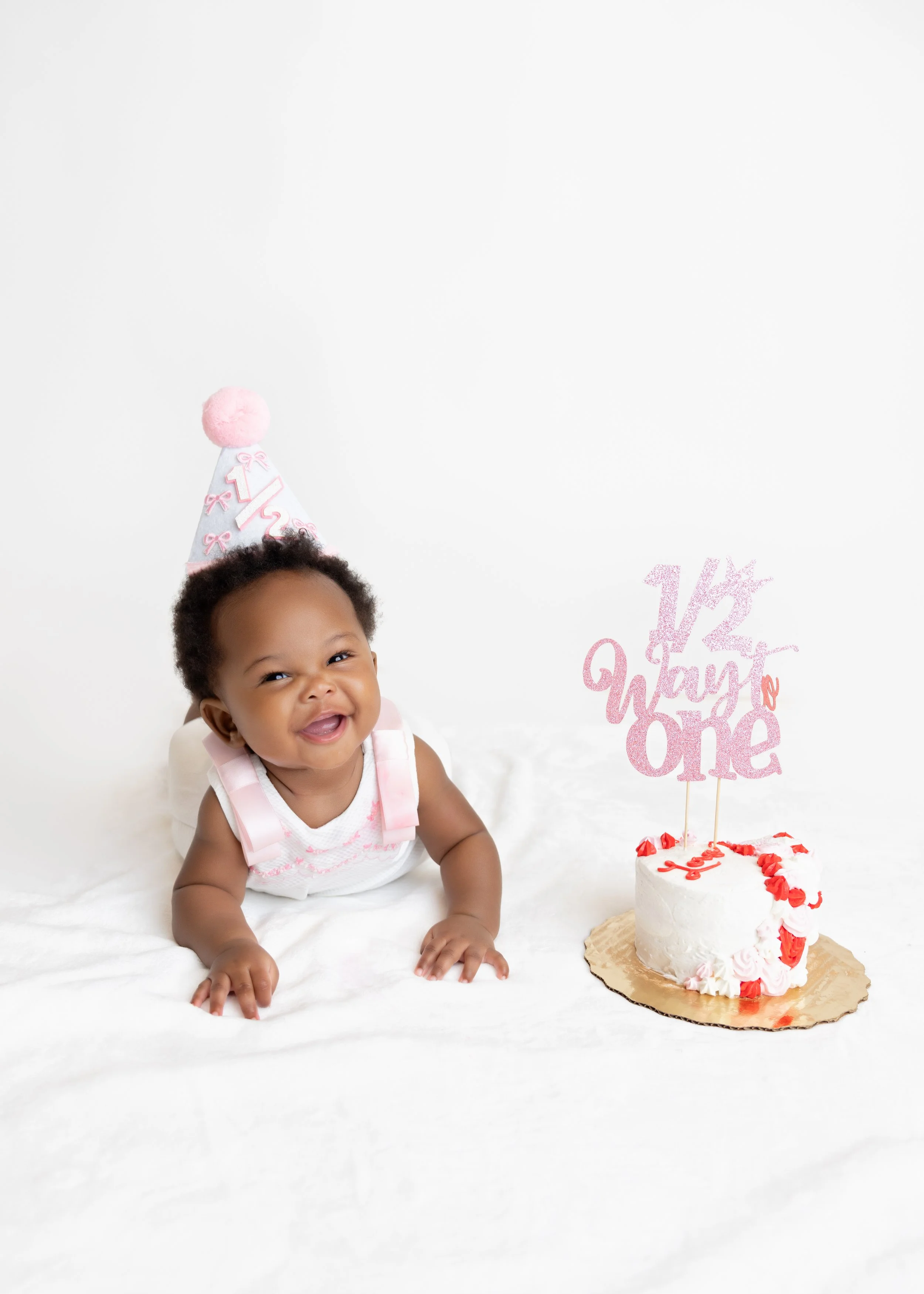 Baby girl celebrating her first birthday with a pink and white cake, wearing a pink and white outfit, and a pink birthday hat with the number 1 on it.