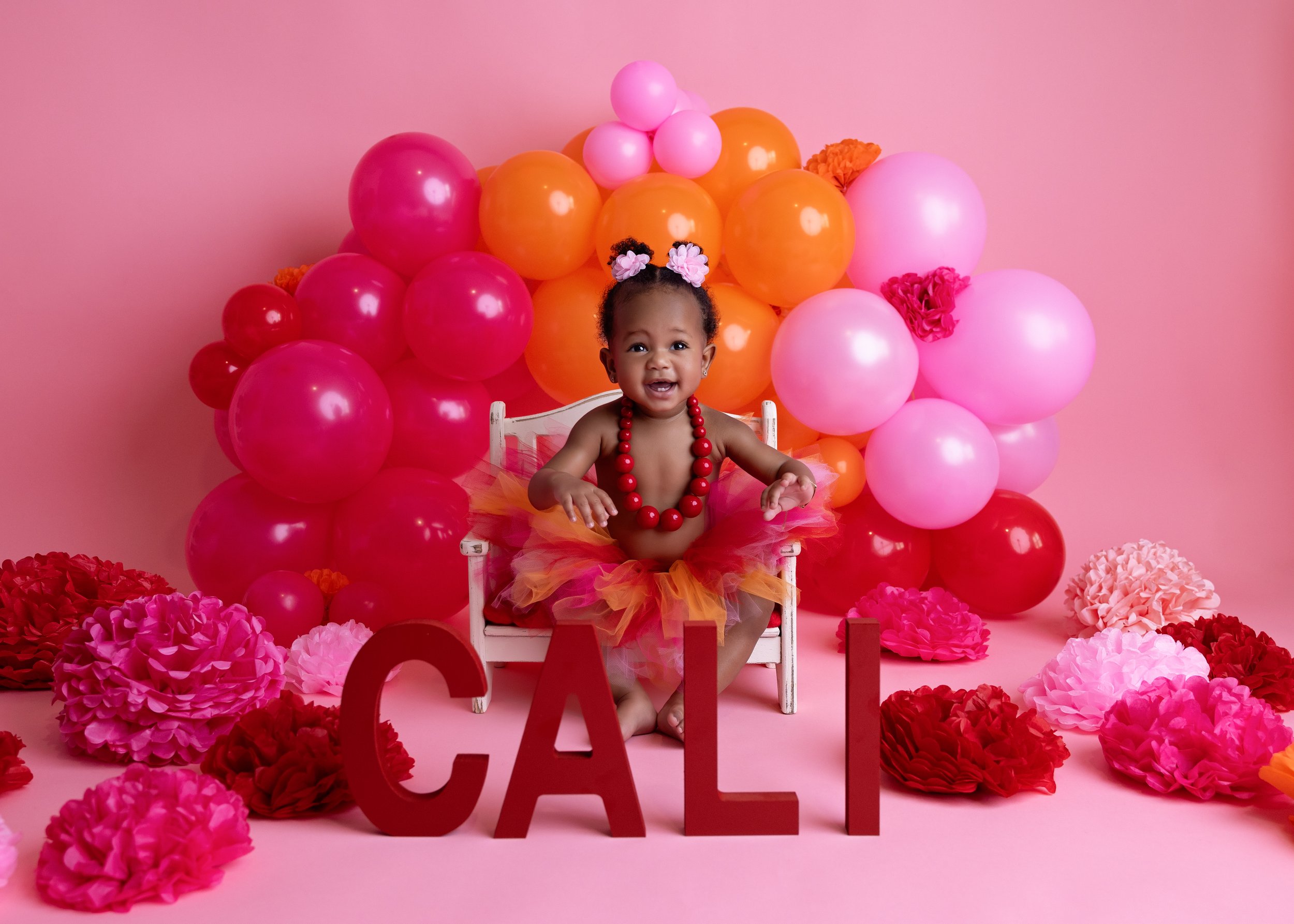 A happy baby girl sitting on a small white chair with pink, red, and orange balloons and flowers around her, with large red letters spelling 'CALI' in front of her.