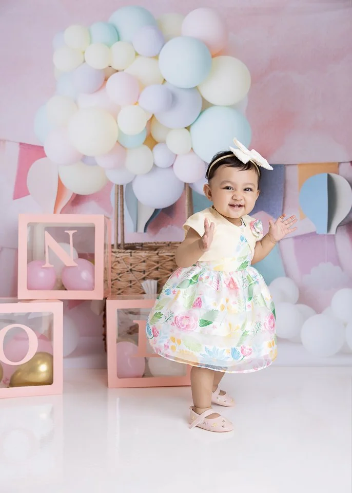 A young girl in a floral dress and pink shoes standing in front of a pastel balloon backdrop and pink boxes spelling out 'N O E,' with some balloons inside the boxes.
