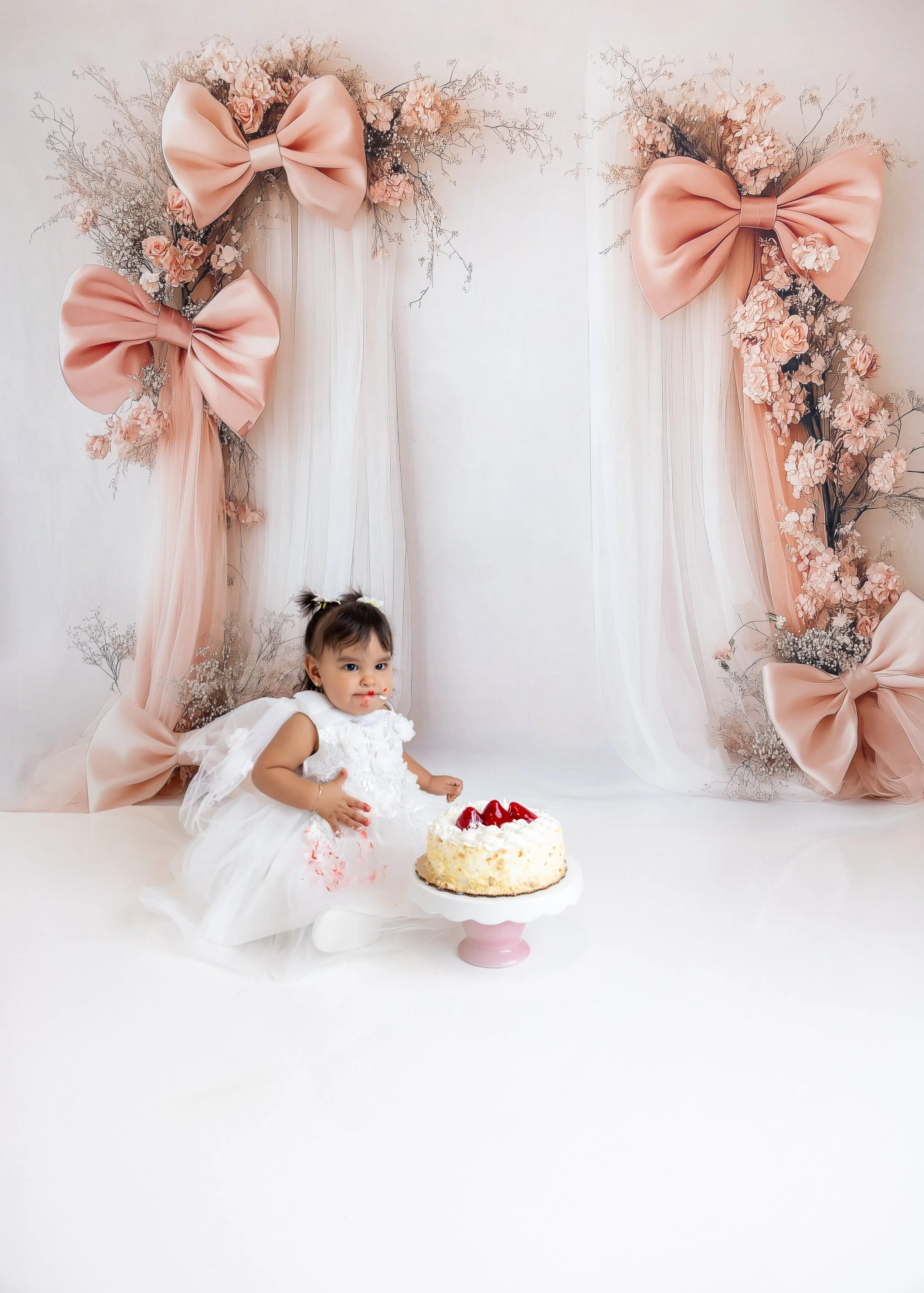 A young girl in a white dress sitting on the floor with a birthday cake decorated with strawberries in front of her, in front of a backdrop with pink bows and flowers.