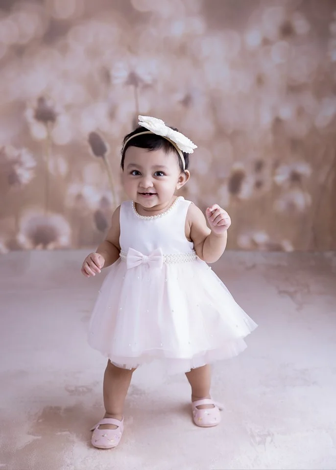 A smiling baby girl in a white dress with a pink bow, wearing a headband with a large flower and pink shoes, standing on a light-colored floor with a floral blurred background.