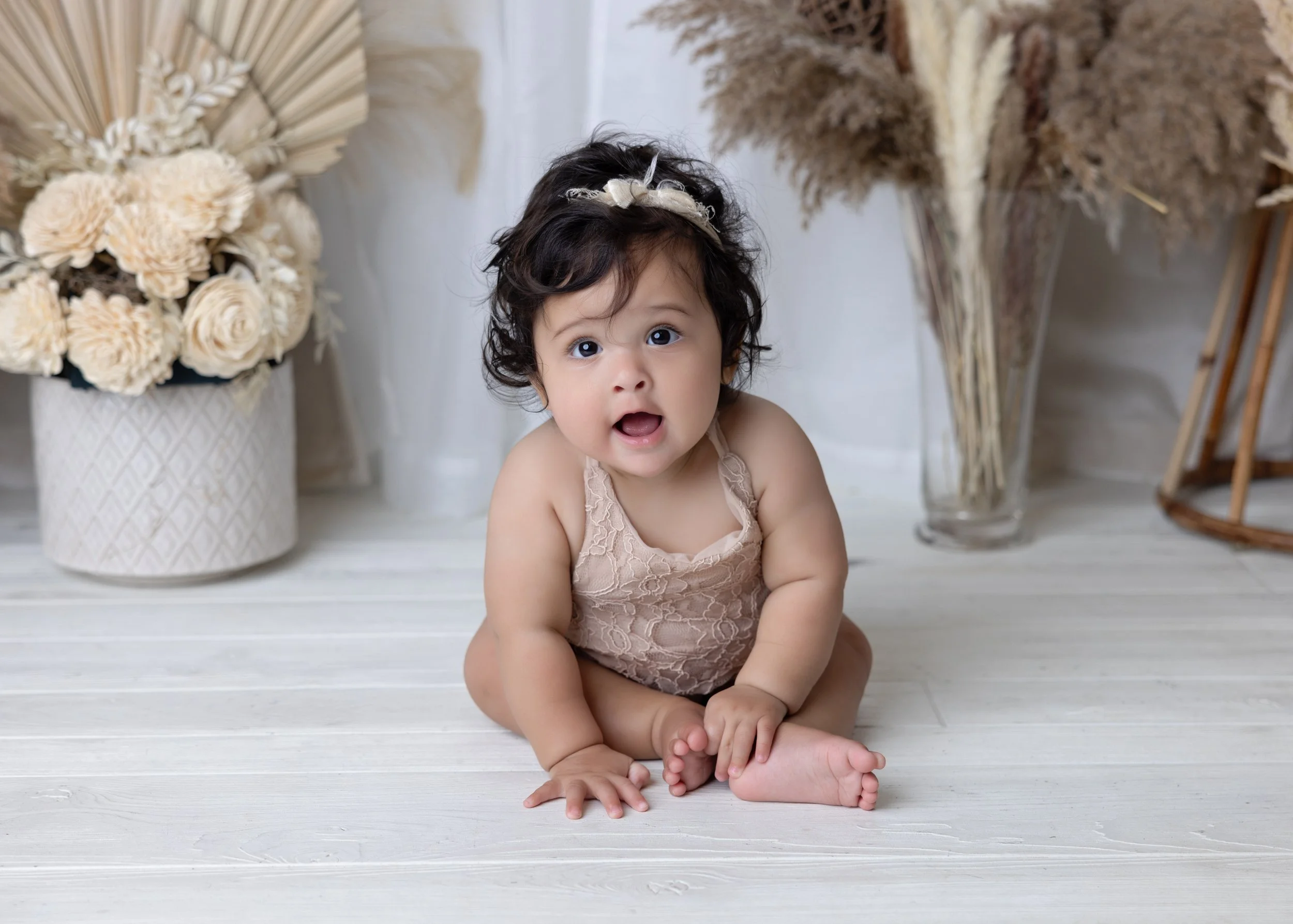 A curious toddler girl with dark curly hair, sitting on a white wooden floor, surrounded by beige and cream-colored flowers and dried plants in vases, wearing a beige lace sleeveless romper and a cream headband with a bow.