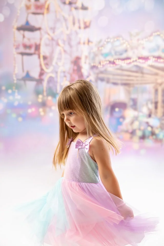 A young girl with long light brown hair wearing a pastel-colored dress with a bow on the shoulder, standing in front of a blurry amusement park or fairground with a Ferris wheel and colorful lights.