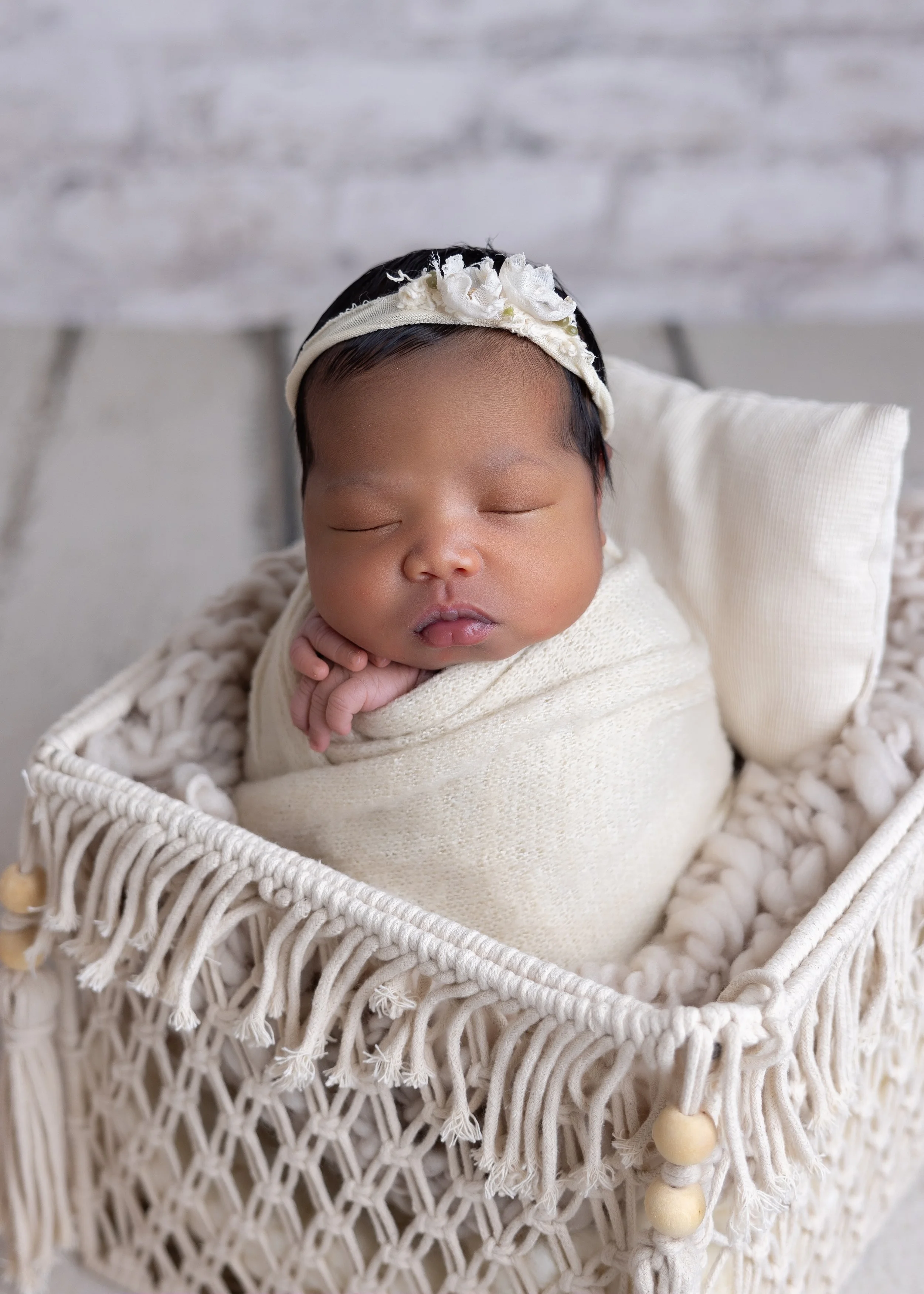 A sleeping baby wrapped in a cream-colored blanket, lying in a woven basket with a fringed edge and beads, with a white pillow behind head and a black headband decorated with white flowers.