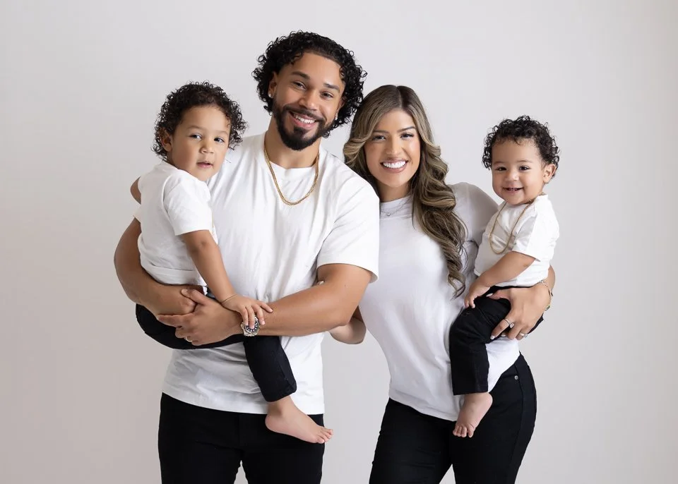 A smiling family of four, consisting of a man, a woman, and two young children, posing together against a plain light gray background.