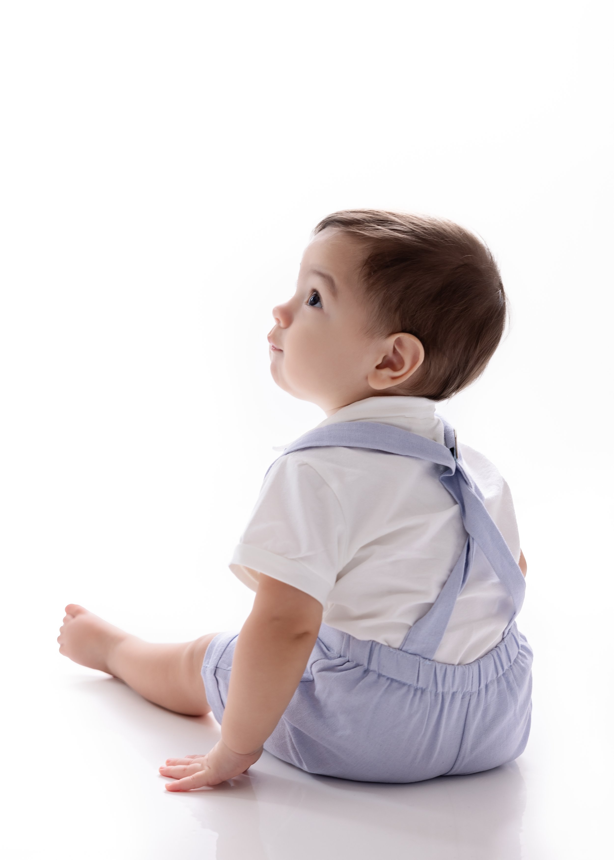 Side view of a young child sitting on the floor, looking upwards with curiosity, wearing a white shirt and light purple shorts with suspenders, against a plain white background.