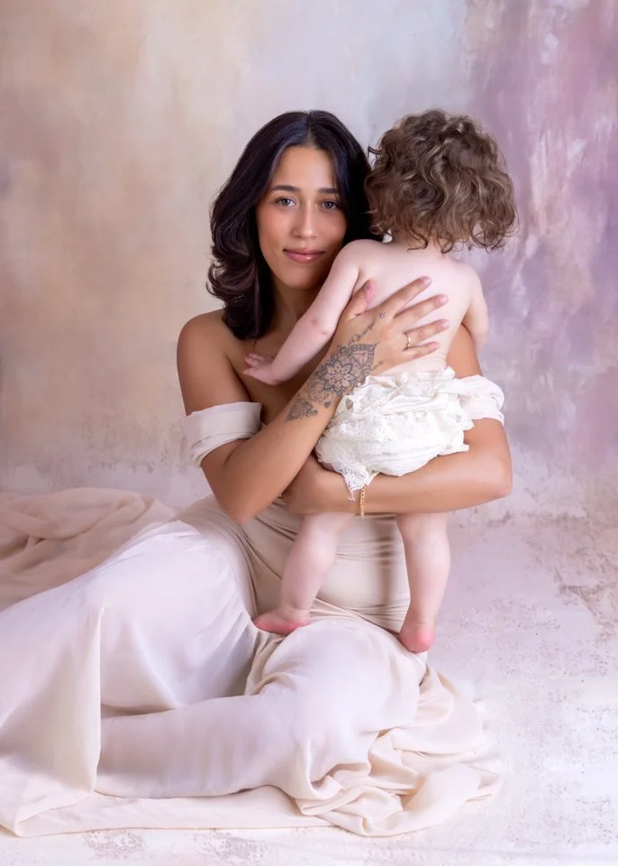 A young woman with dark hair and tattoos on her arm holding a small child with curly hair, sitting on a beige fabric against a textured pastel background.