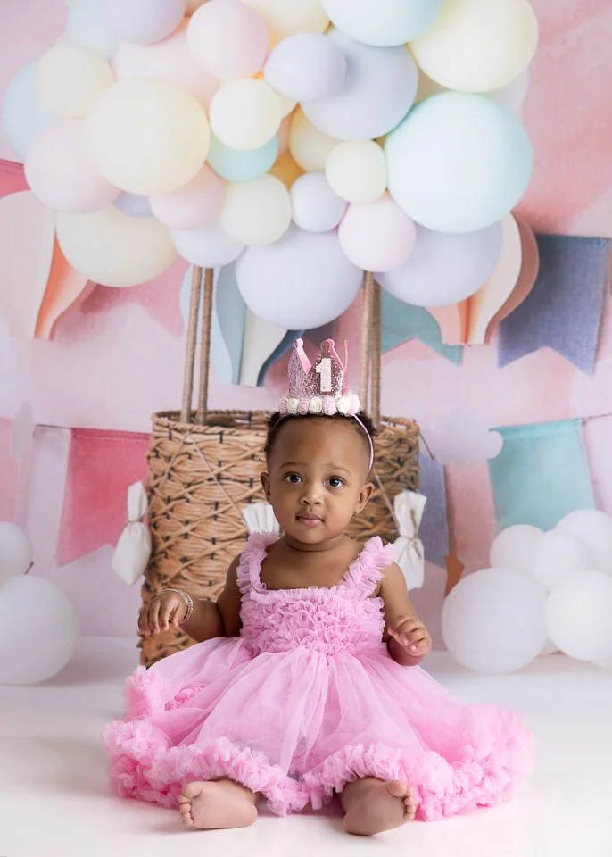 Baby girl sitting on the floor in pink dress with birthday crown, background decorated with pastel balloons and paper banners for first birthday celebration.