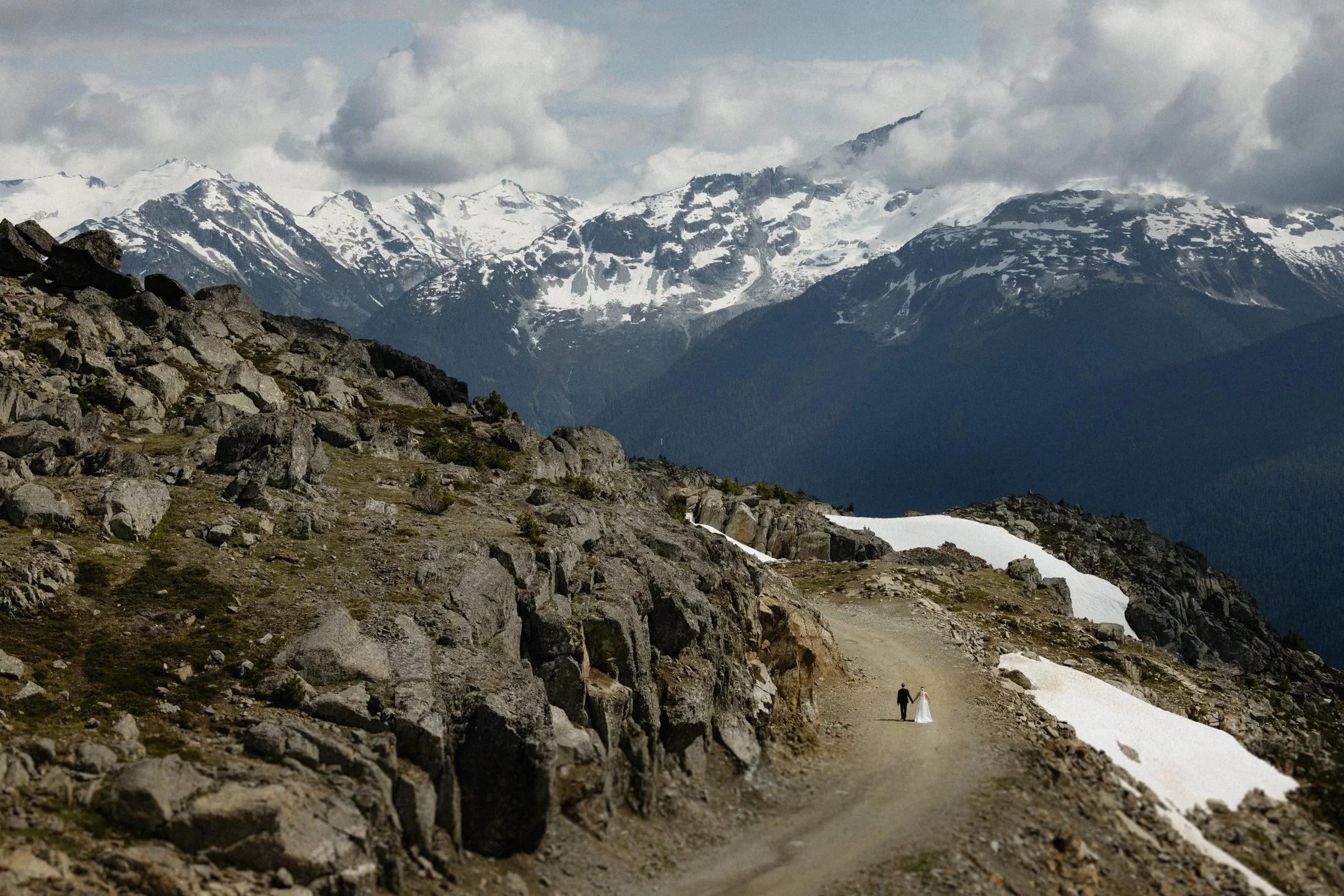 A far away image of a couple walking hand in hand down a gravel road among the rocks and mountains of Whistler Mountain