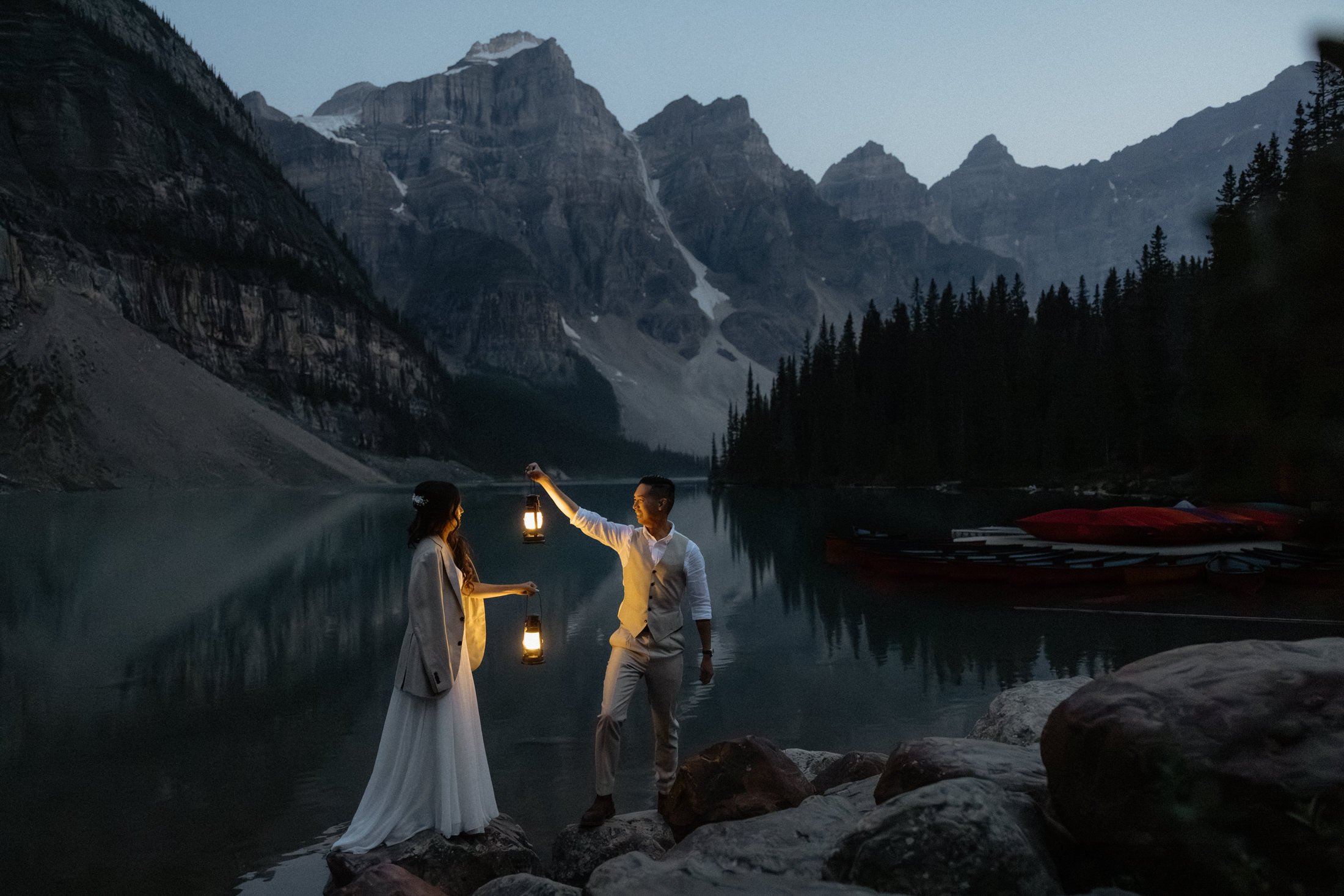 A newly married couple stands at the edge of moraine lake at dusk holding lanterns to illuminate themselves in banff national park