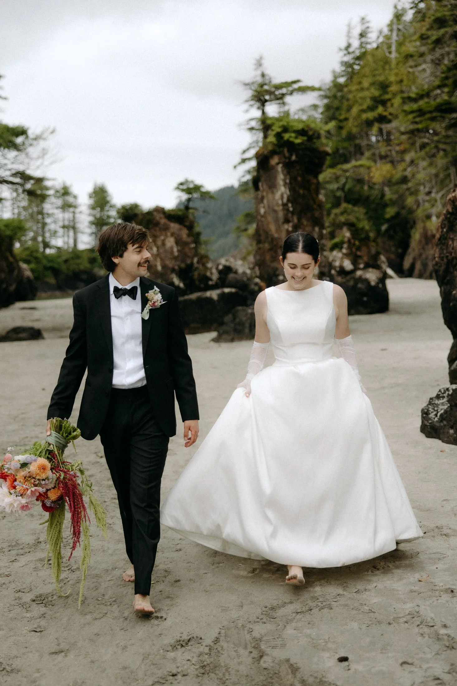 An elopement couple walks hand in hand on the beach among the sea stacks of san josef bay in the background in cape scott provincial park