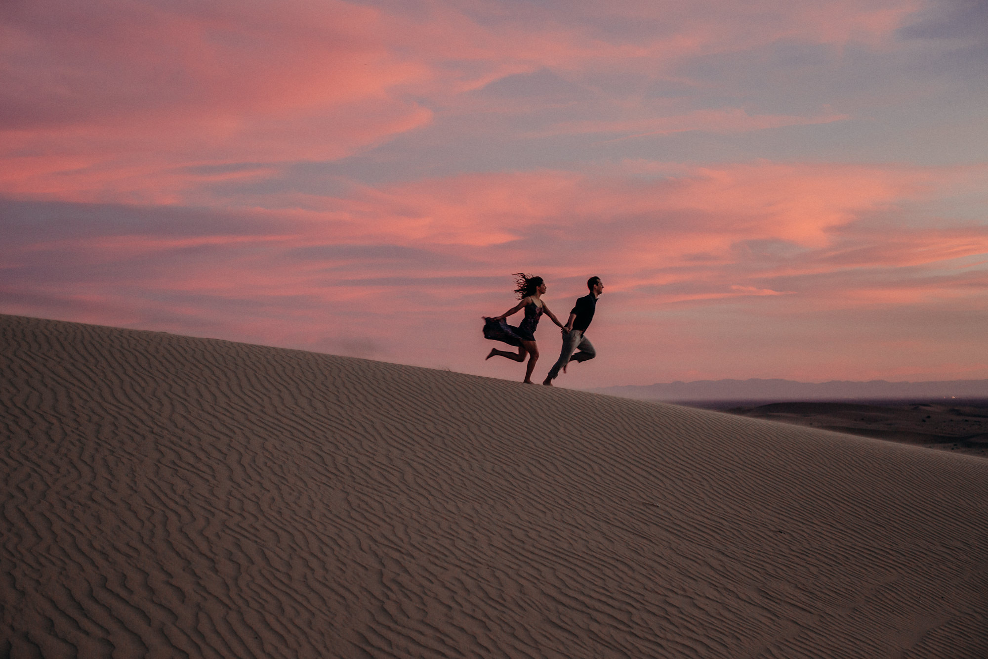 A silhouetted couple running down the sand dunes with a cotton candy sky behind them at the Glamis Imperial Sand Dunes. Photographed by Candice Marie Photography.