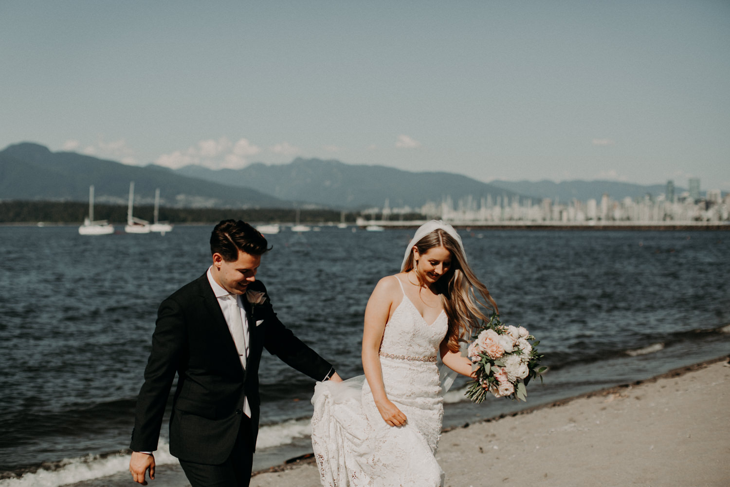 The bride and groom walk on the sand by the water at Jericho Beach in Vancouver, BC.