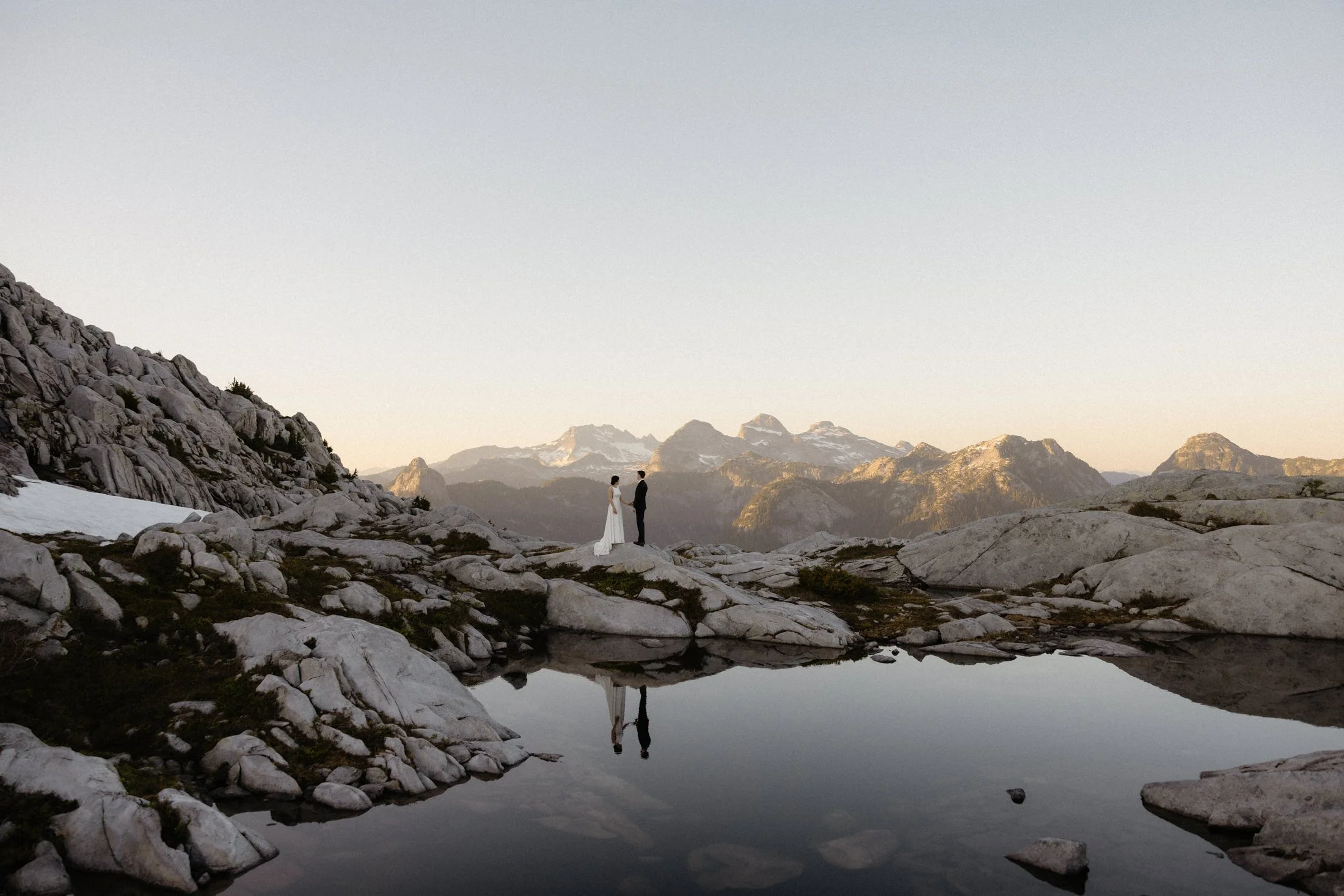an elopement couple stands on some rocks behind a pond with their reflection and mountains in the background at sunset, made possible by bc helicopters