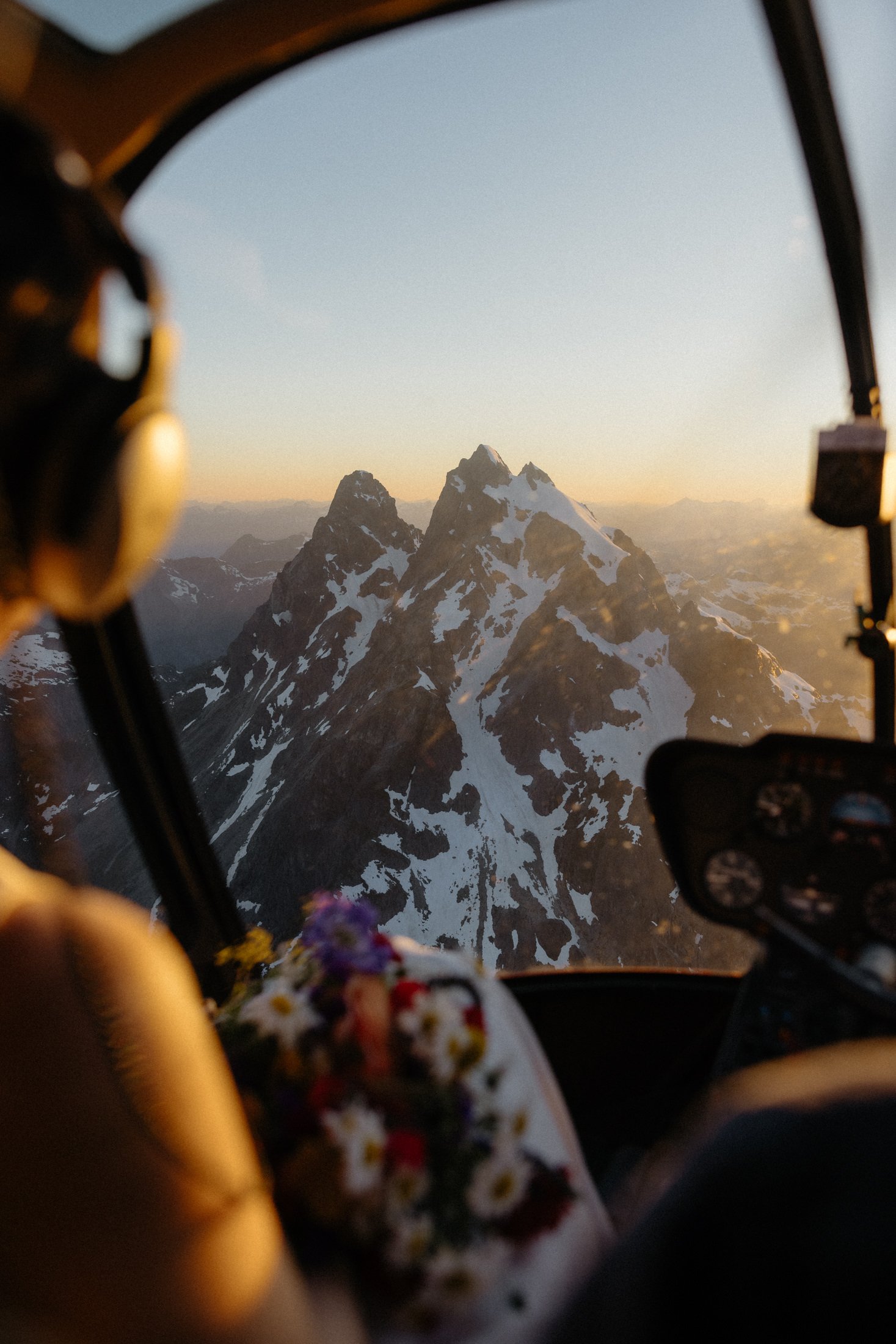 A view from a helicopter, over a brides shoulder, looking at a snow capped mountain at sunset