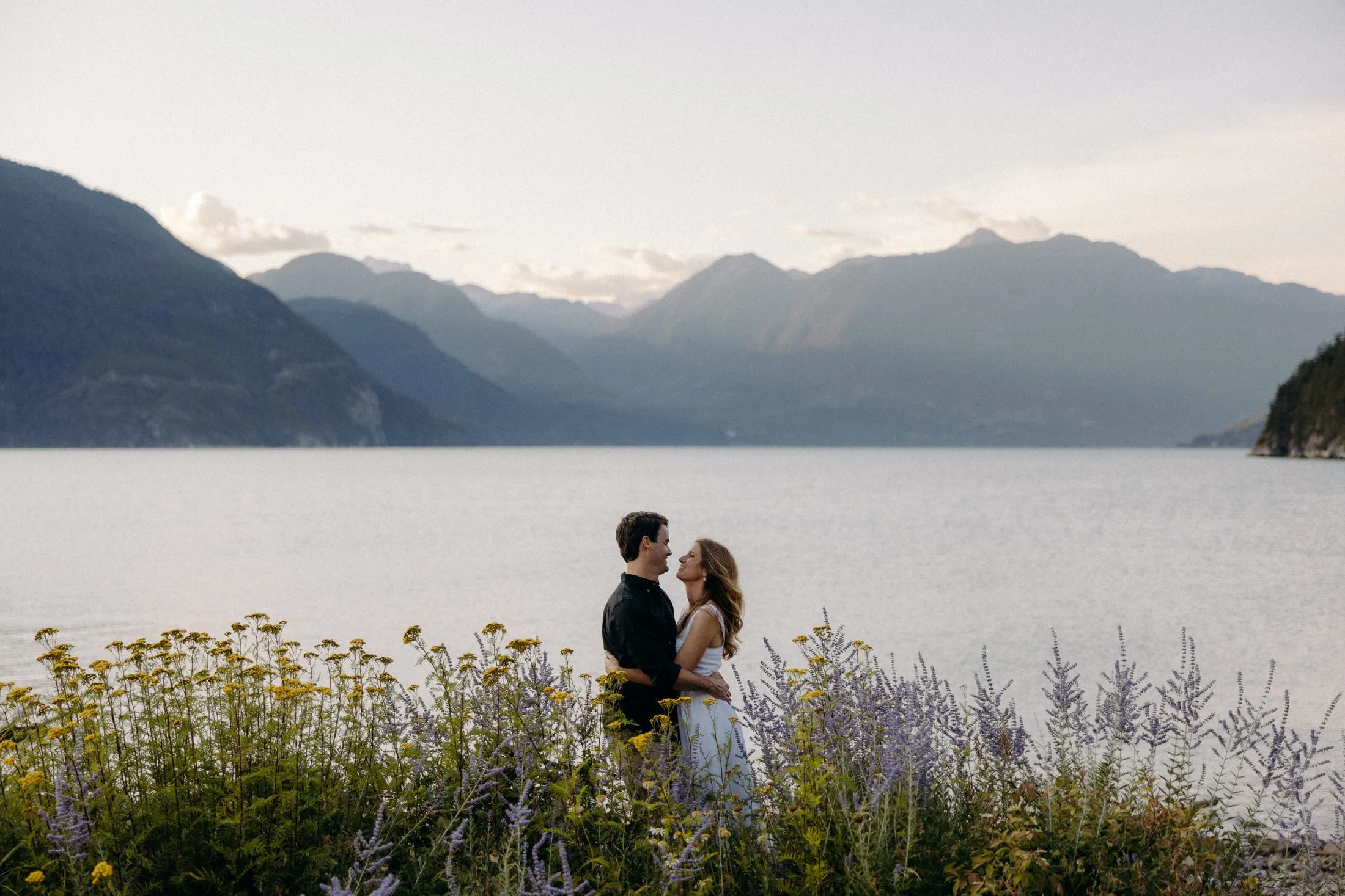 A couple embraces and looks at each other among purple and yellow flowers with the ocean and mountains in the background at oliver's landing in Squamish, BC