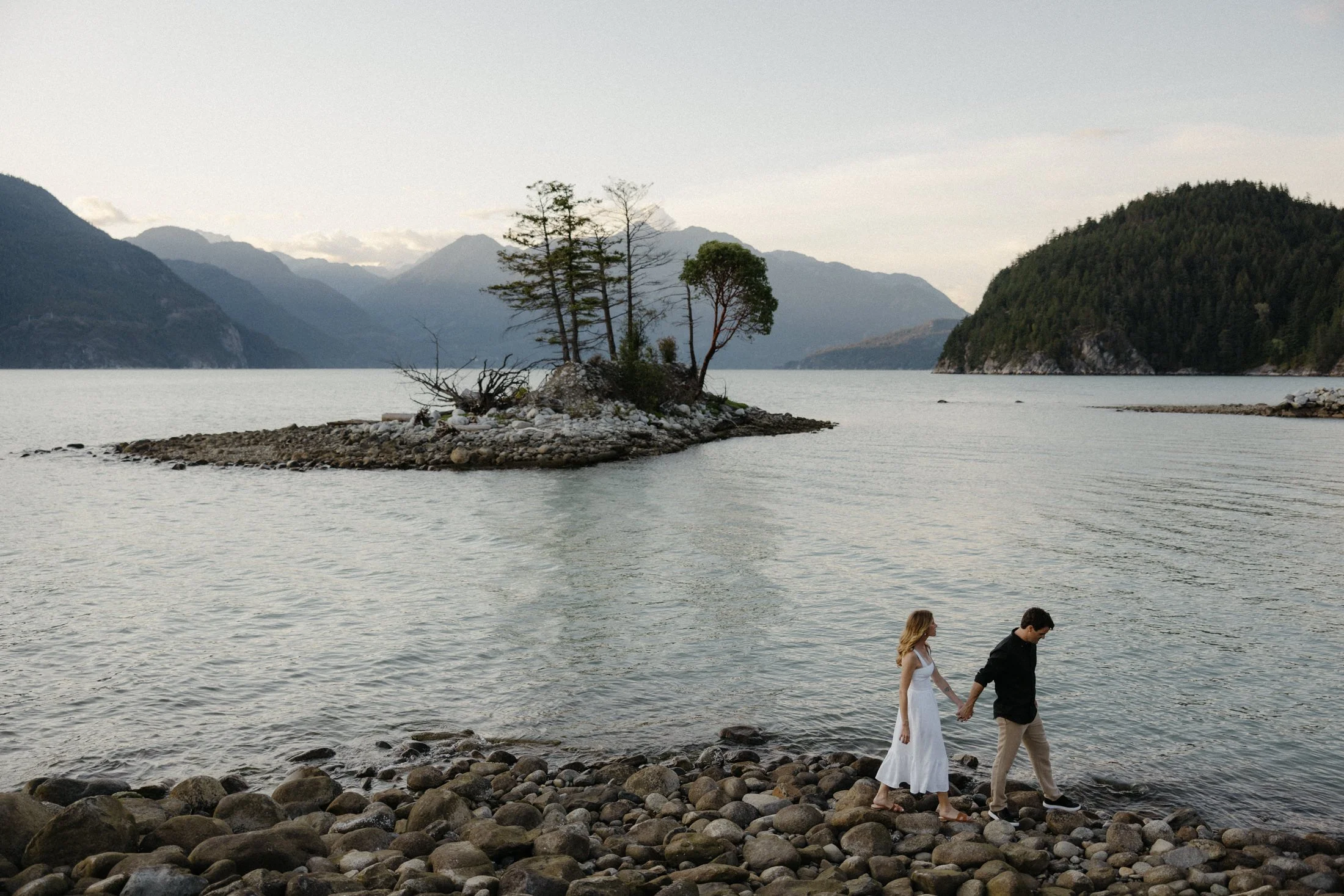 A couple walks hand in hand on the rocks by the ocean with a small rocky island and mountains in the background at oliver's landing near Squamish, BC