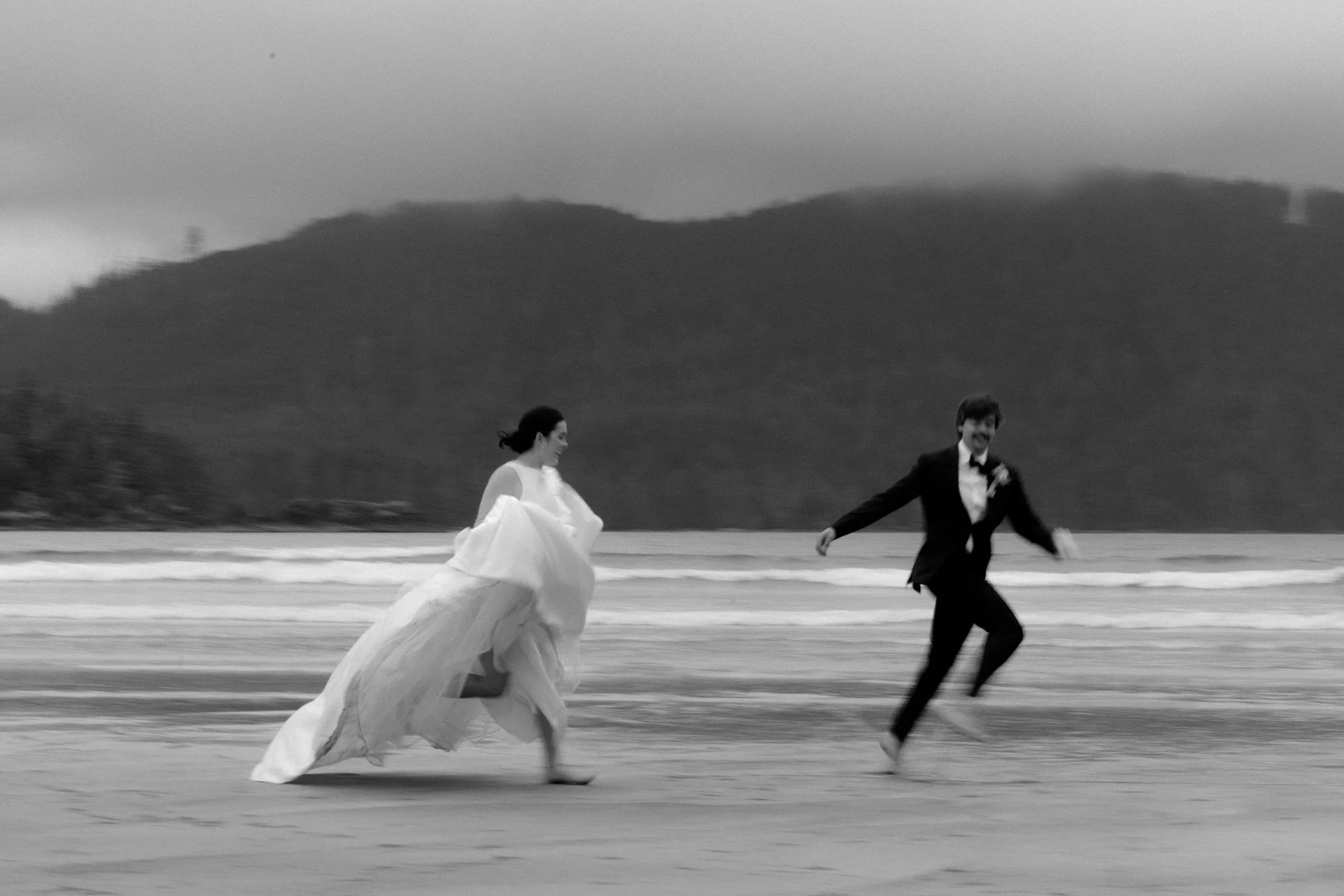 A semi blurry, black and white image of an elopement couple running down the beach, chasing each other at san josef bay in cape scott provincial park