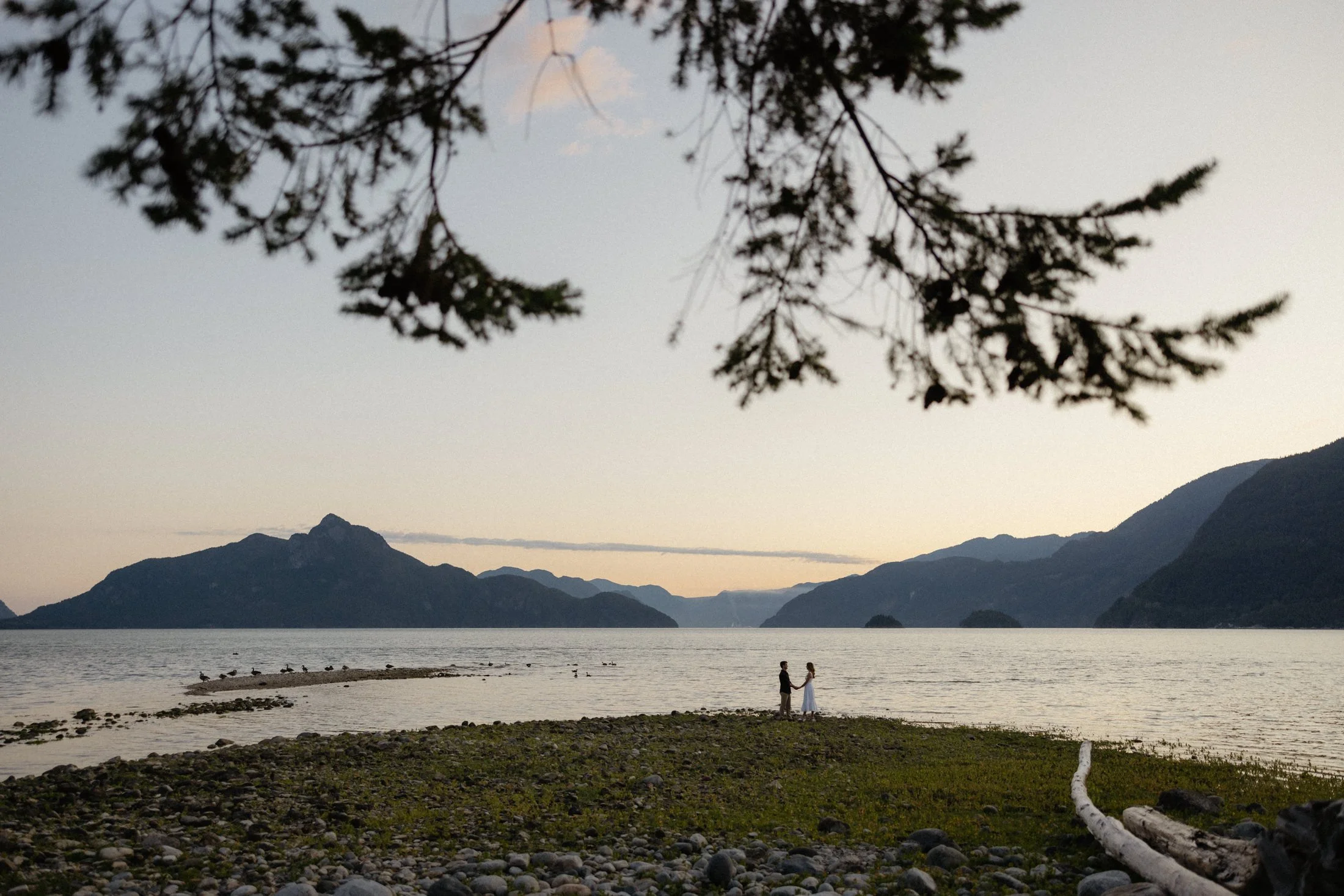A couple stands distantly on the edge of a rocky beach at sunset with the ocean and mountains in the background at oliver's landing in Squamish, BC