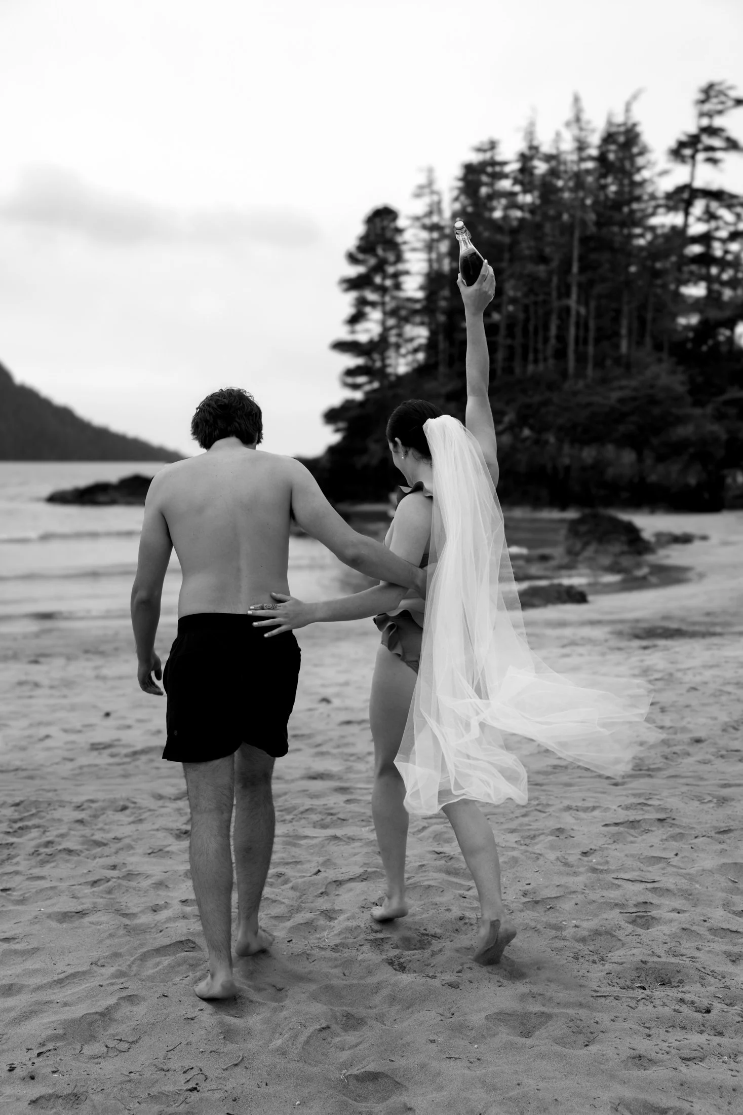 a newly married couple with bathing suits and the bride wearing a veil, walks towards the ocean on the beach, about to take a swim in san josef bay in cape scott provincial park
