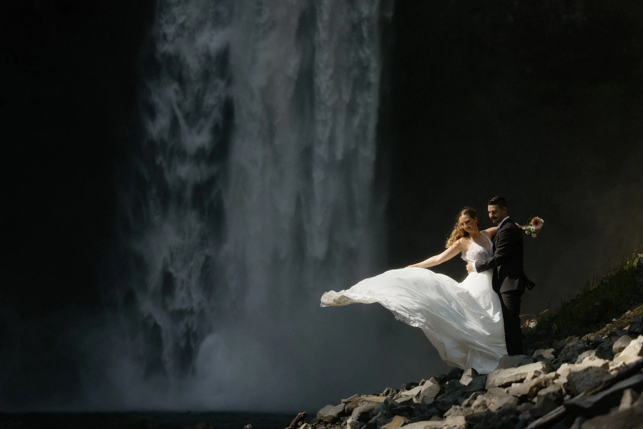 An elopement couple embraces as the bride throws her dress and it catches in the wind, standing in the sunlight in front of a large waterfall in whistler, bc