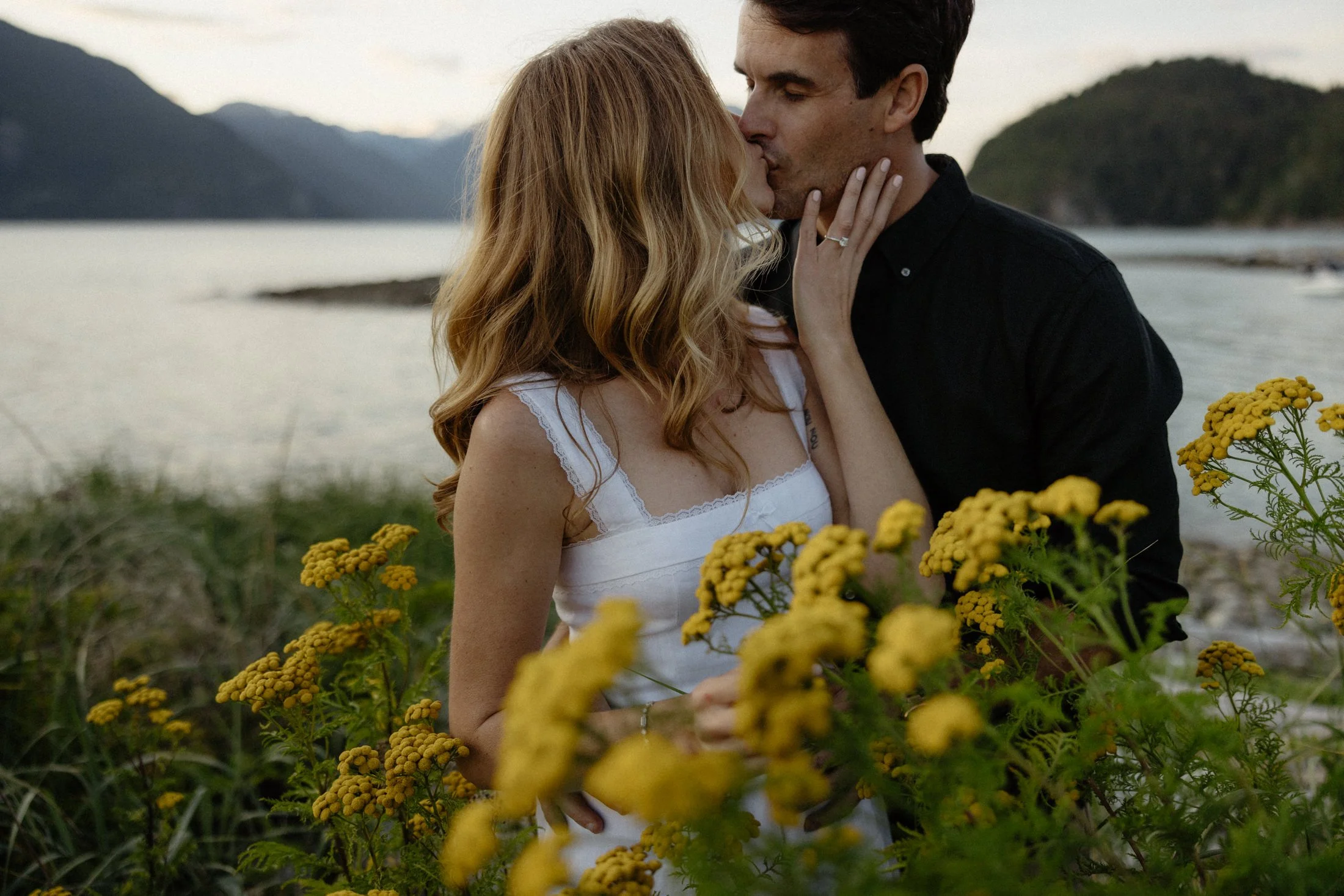 A couple kisses among yellow flowers with the ocean and mountains in the background at oliver's landing in squamish, BC