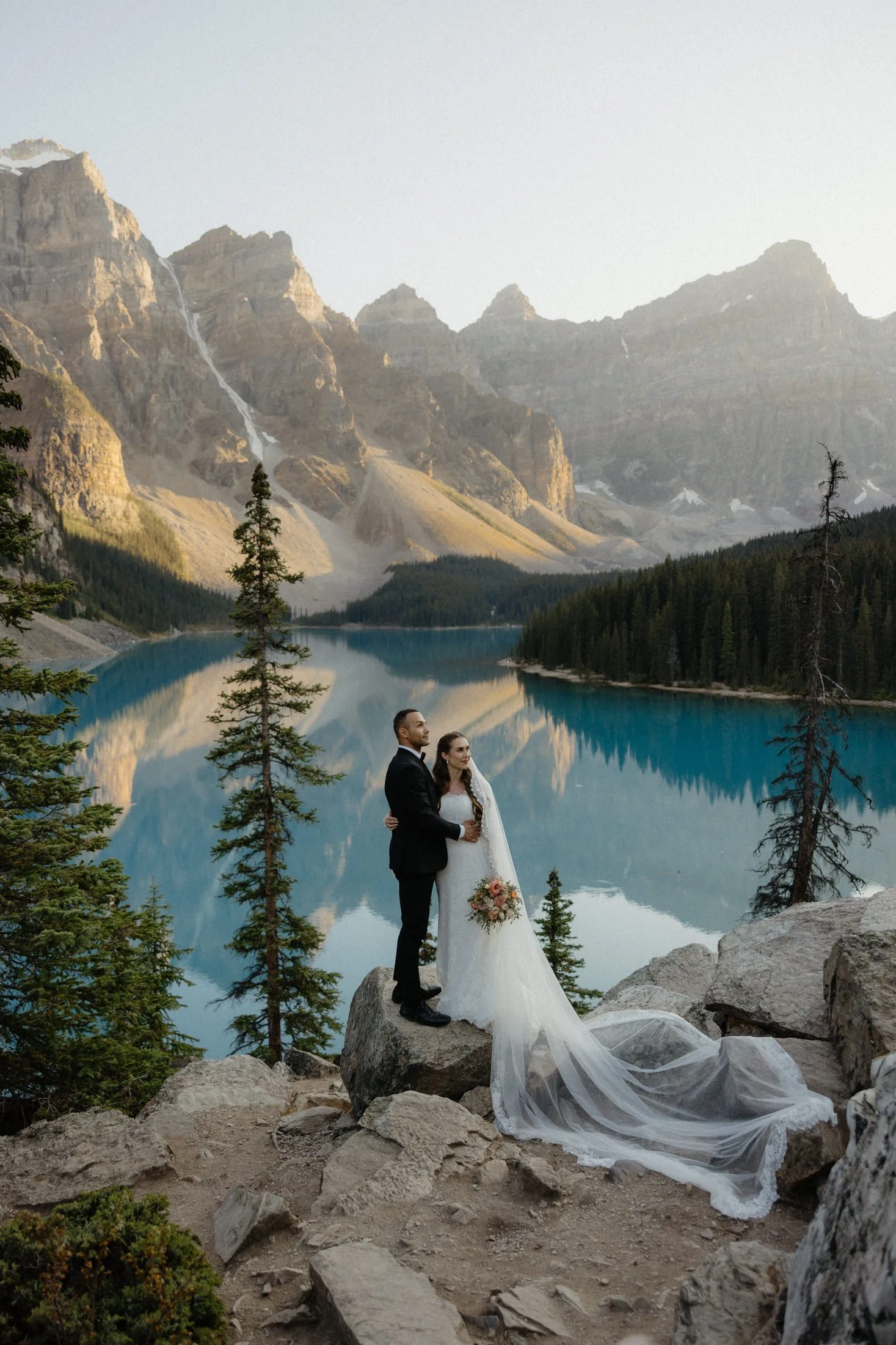 An elopement couple stands embracing on a rock with the bride's veil draped over the rocks with the turquoise blue waters of Moraine lake and mountains in the background in Banff national park