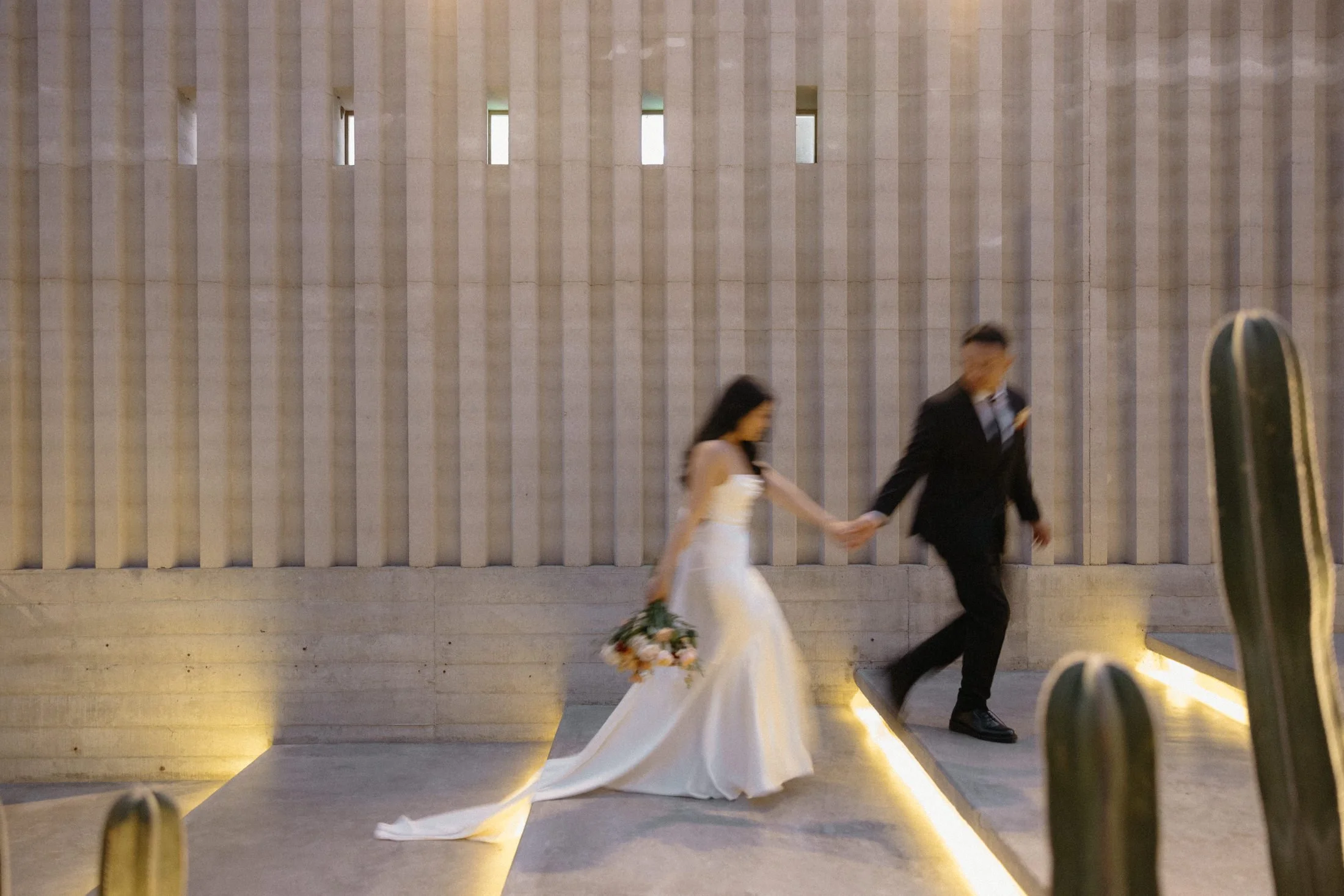 a wedding elopement couple walks up the steps in a blurry motion in front of the concrete wall near the acre restaurant and cocktail bar in Cabo, Mexico