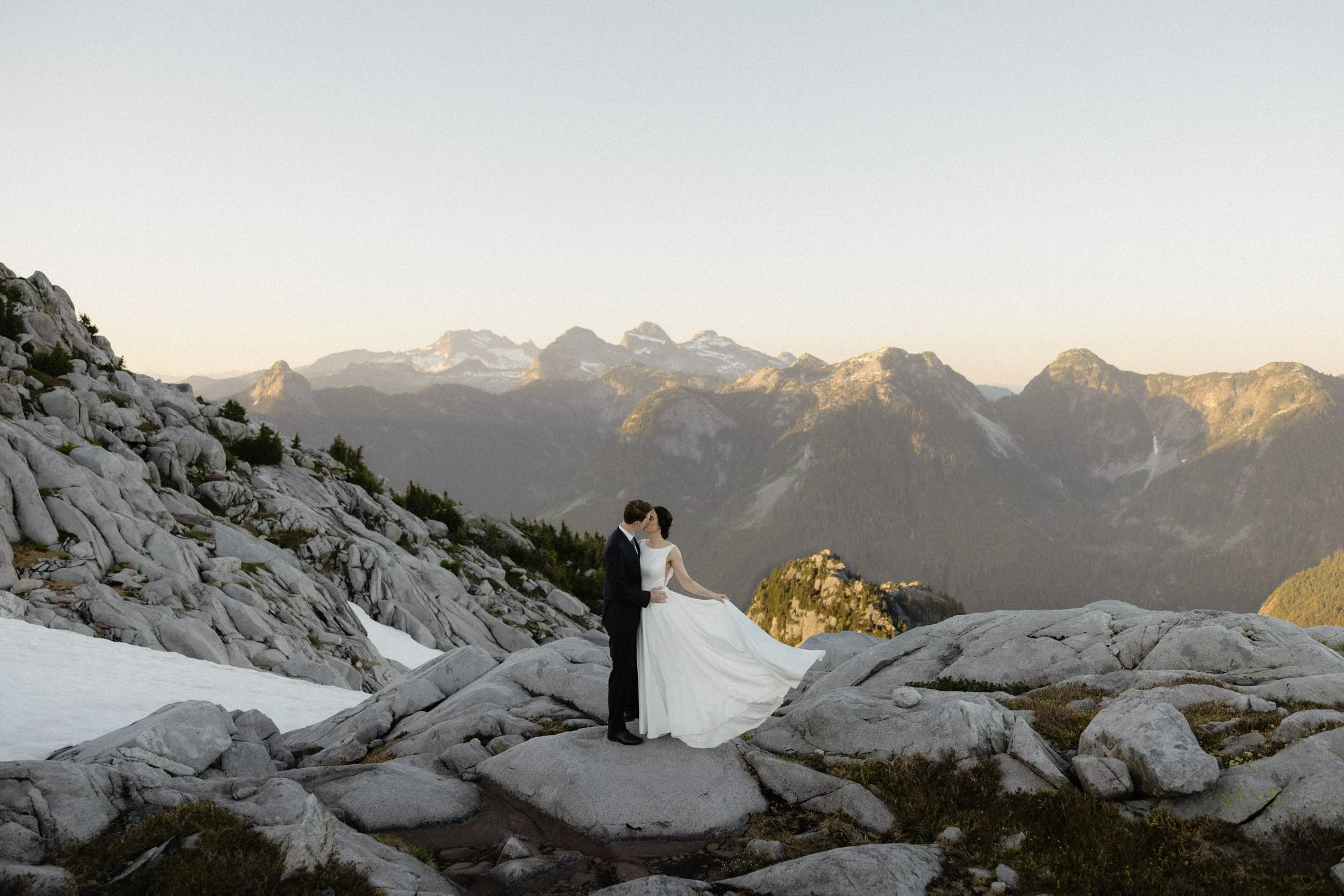 a couple kisses on a rock with mountains in the background at sunset, made possible by bc helicopters