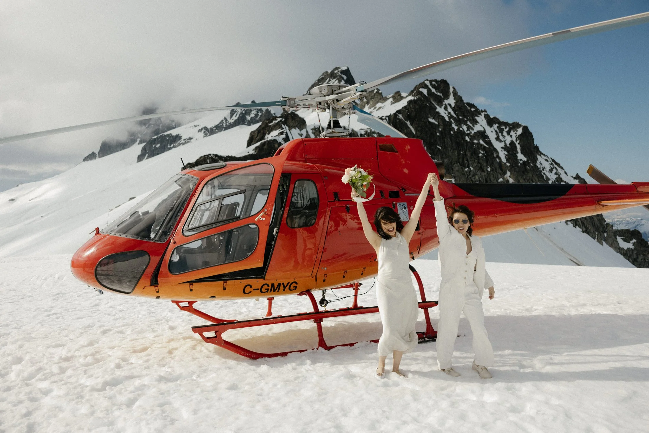 A lesbian couple exits a red helicopter that has just landed on a glacier, holding hands and raising them in the air to cheer, in Squamish, BC