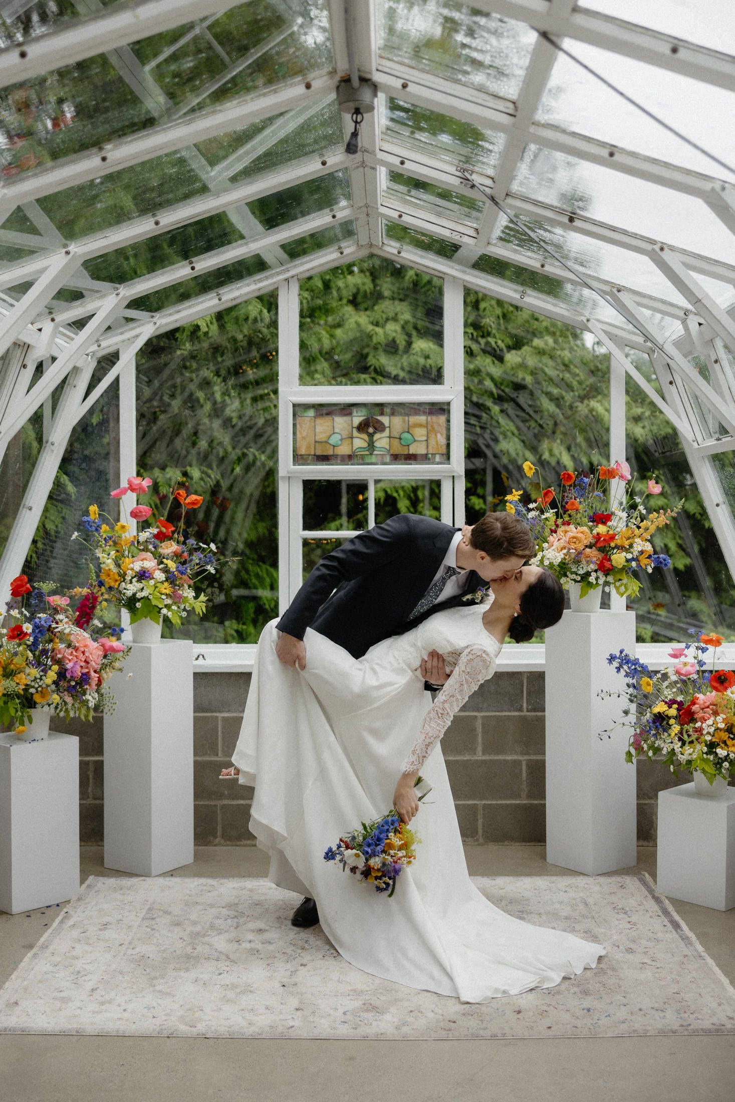 a groom dips his bride to kiss her on a rug, surrounded by floral pillars in the greenhouse ceremony space on a rainy day