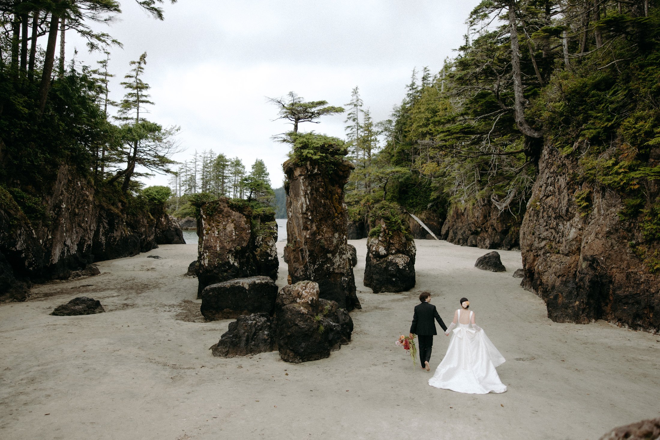 An elopement couple walks hand in hand on the beach among the sea stacks of san josef bay in the background in cape scott provincial park
